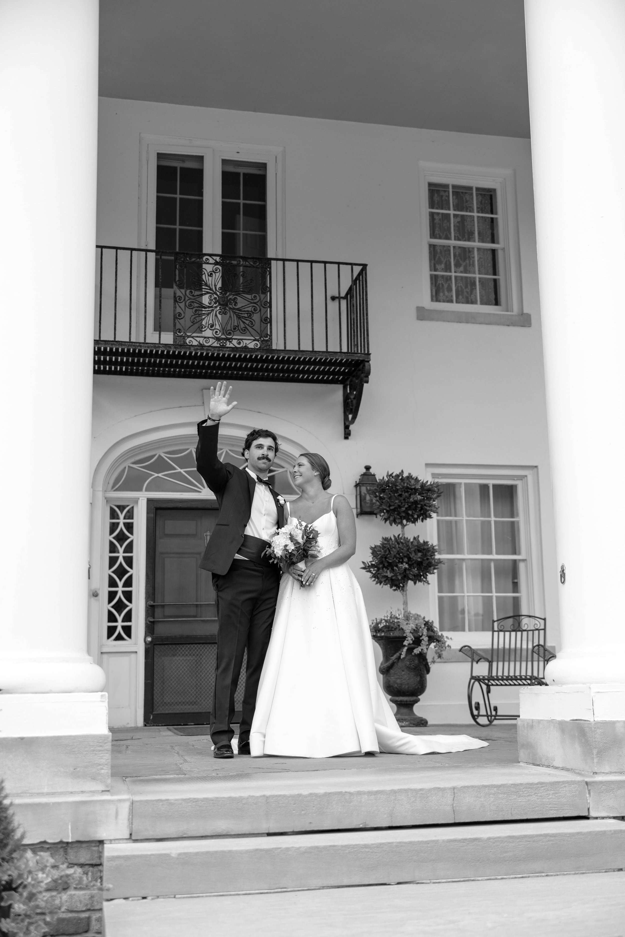A black and white photo of a newly married couple standing on the steps of a large house. The man is dressed in a tuxedo, waving, and the woman is wearing a wedding dress, holding a bouquet, smiling and looking at the man.