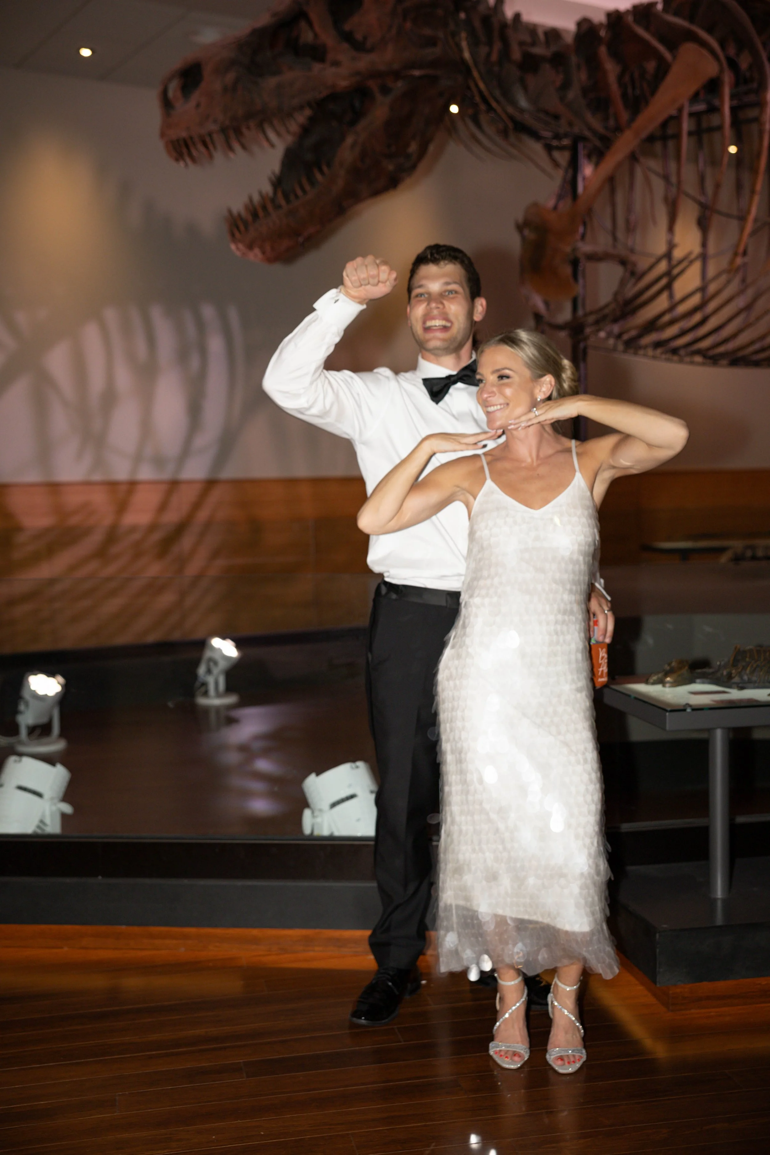 A man and woman dancing together at an event with a large dinosaur skeleton in the background. The man is wearing a tuxedo with a bow tie, and the woman is in a white dress and high heels.