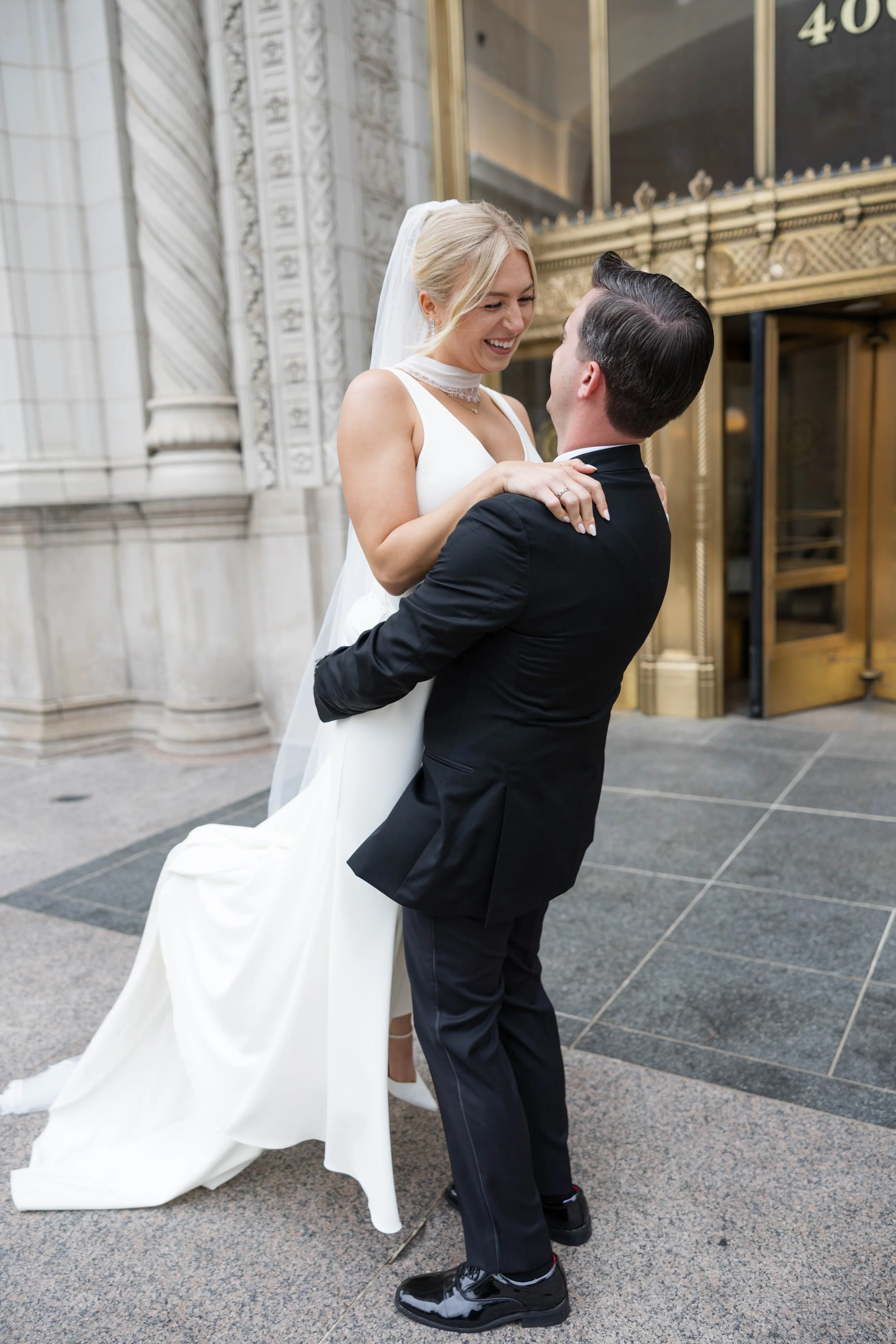 A man in a tuxedo lifting a woman in a wedding dress outside a building with ornate architectural details and, a glass door with gold trim.