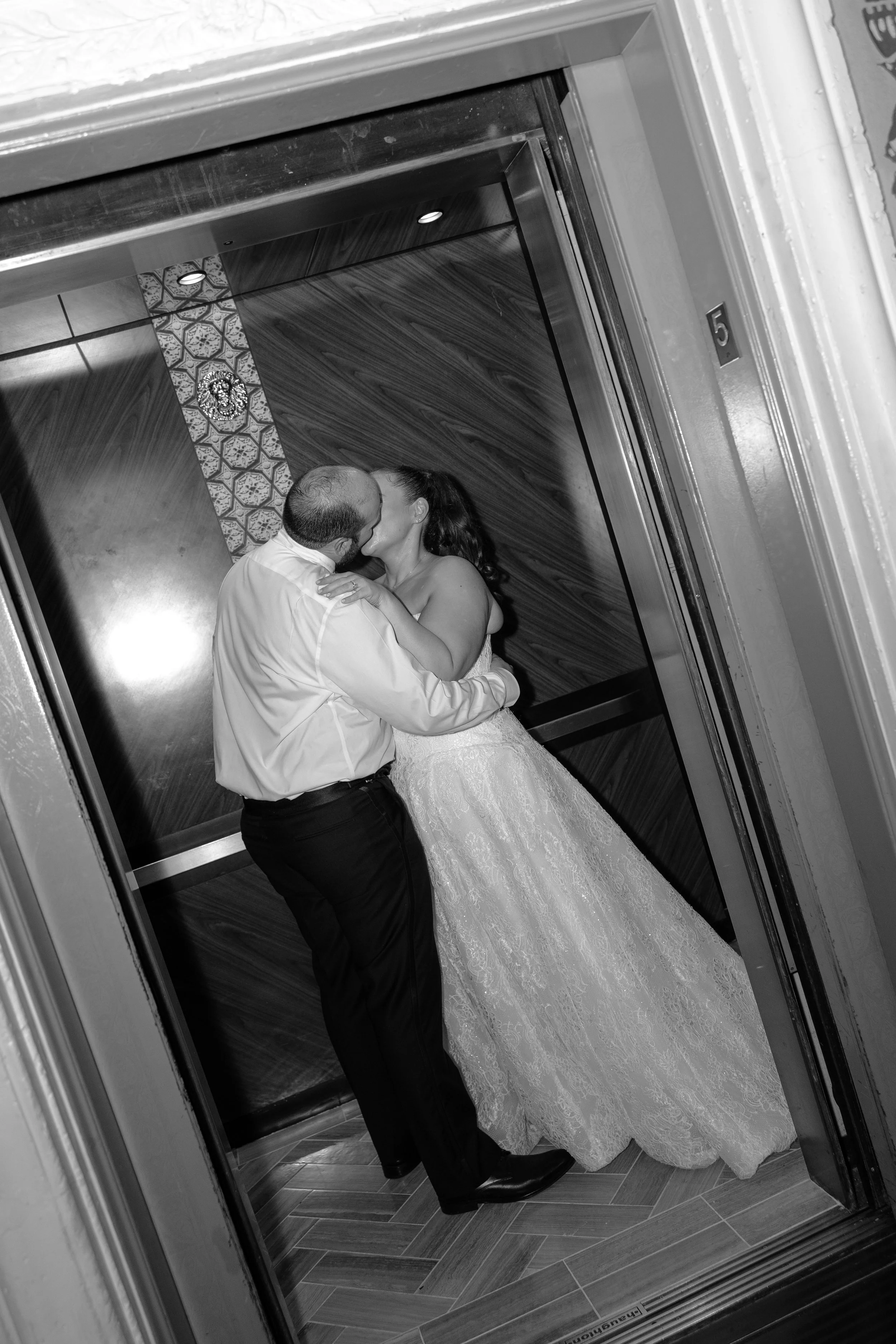 A man and woman share a kiss in an elevator. The man is wearing a light-colored shirt and dark pants, while the woman wears a wedding dress. The elevator has decorative wood paneling and a patterned wall. The photo is in black and white.