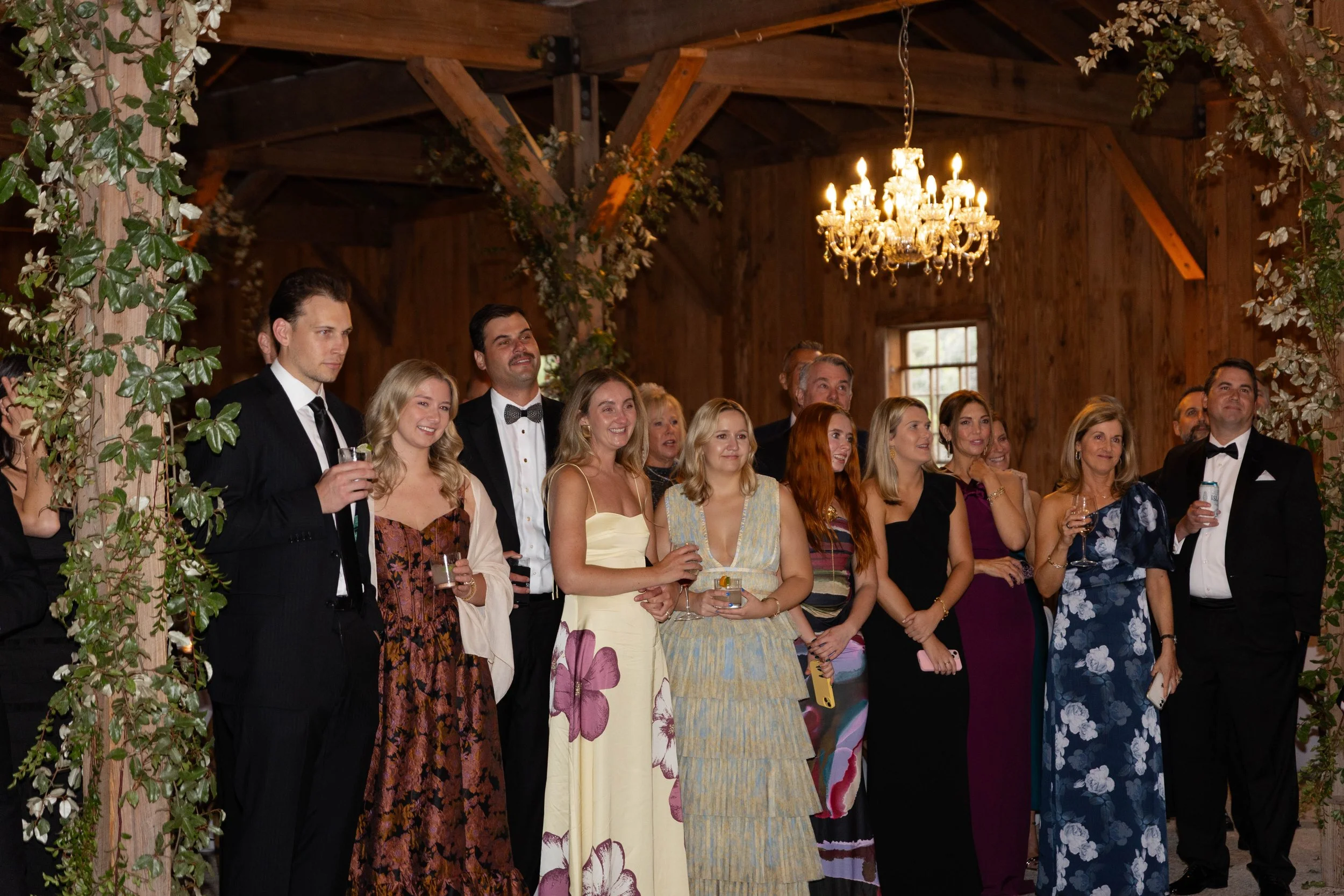 Group of well-dressed people at a celebration or wedding reception in a rustic wooden venue with a chandelier and floral decorations.