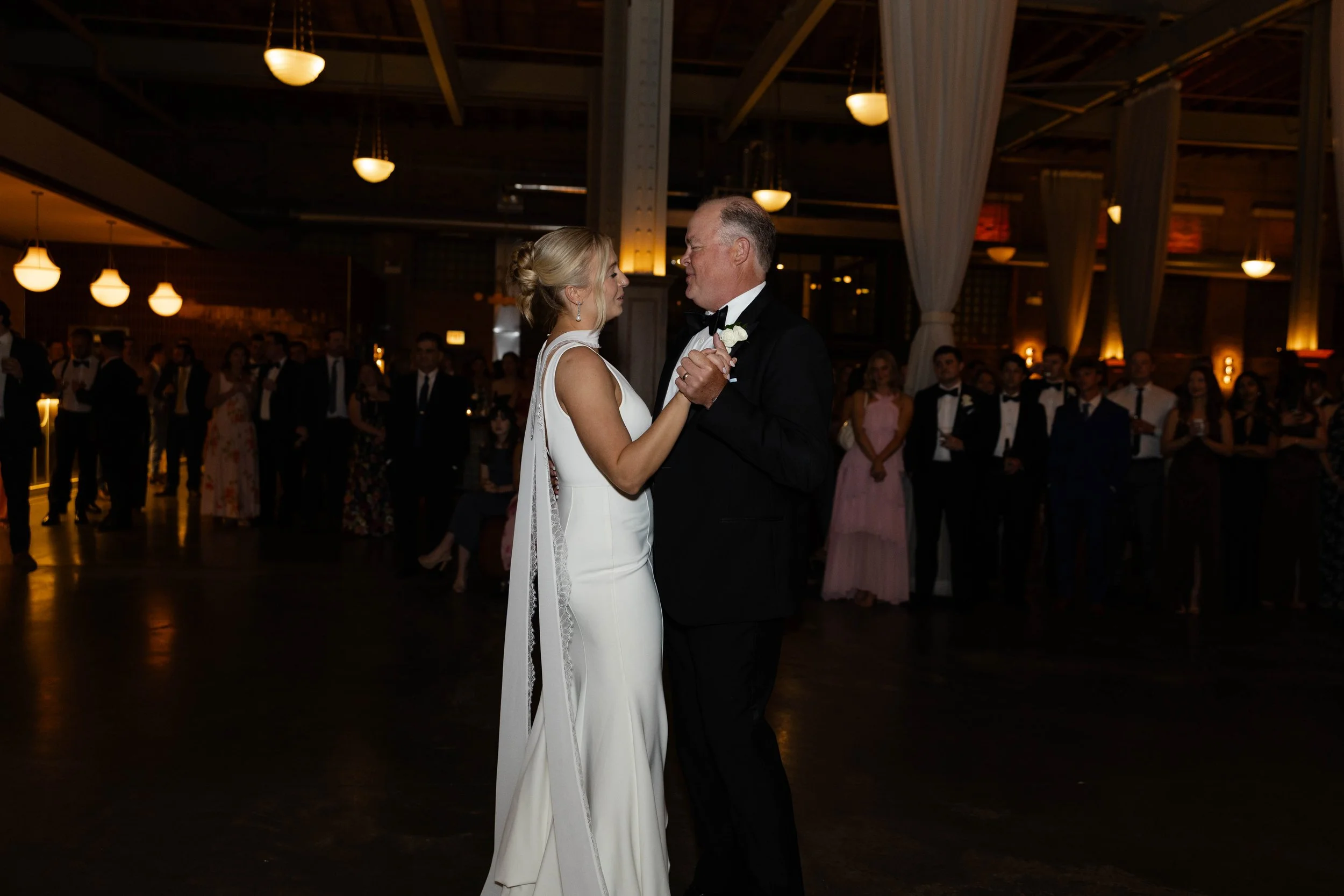 A bride and groom are sharing their first dance at a wedding reception, with guests watching in the background under warm, ambient lighting.