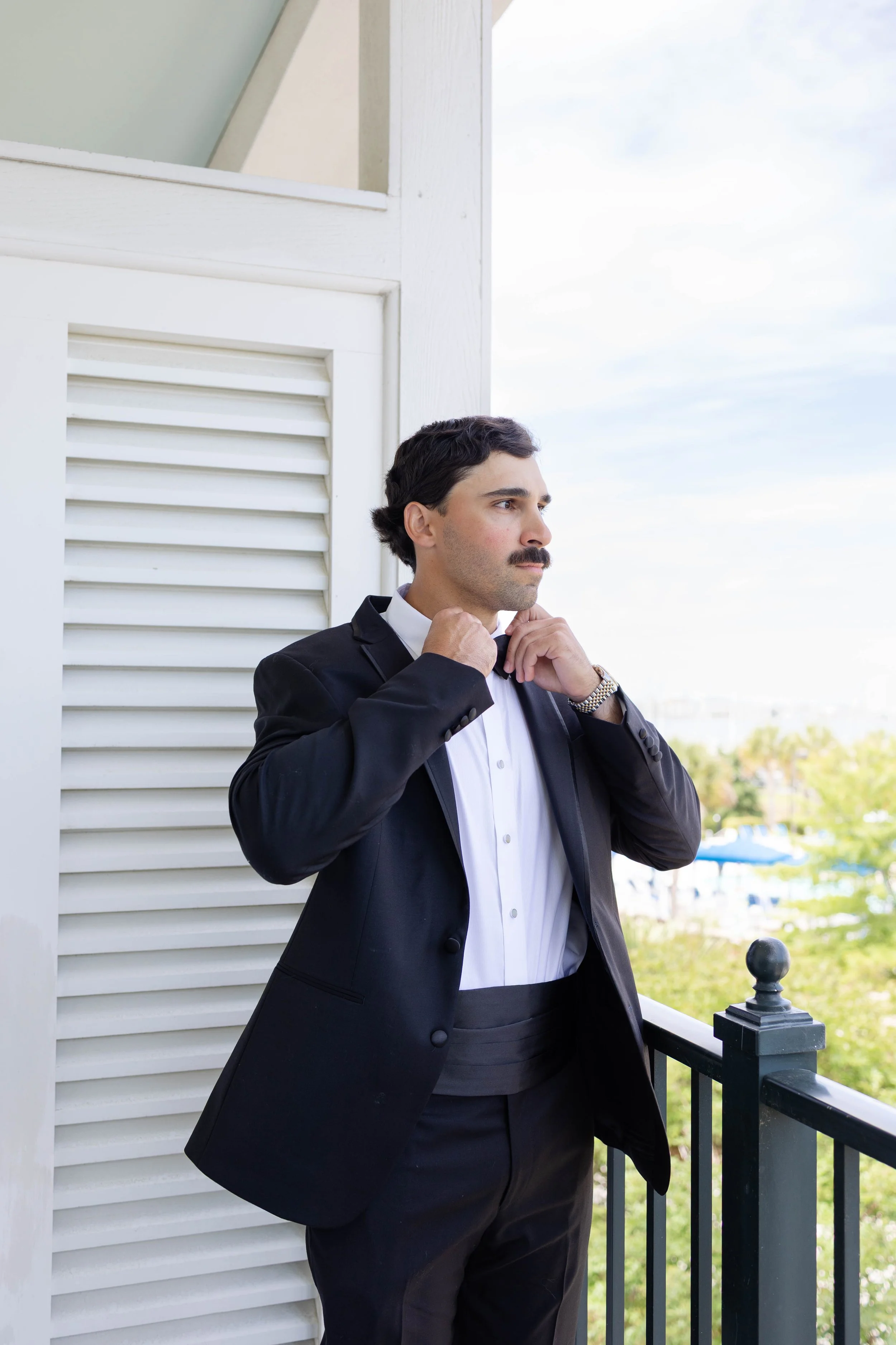 A man with dark hair and a mustache adjusting his bow tie on a balcony, dressed in a black tuxedo with a white shirt, against a background of blue sky and trees.