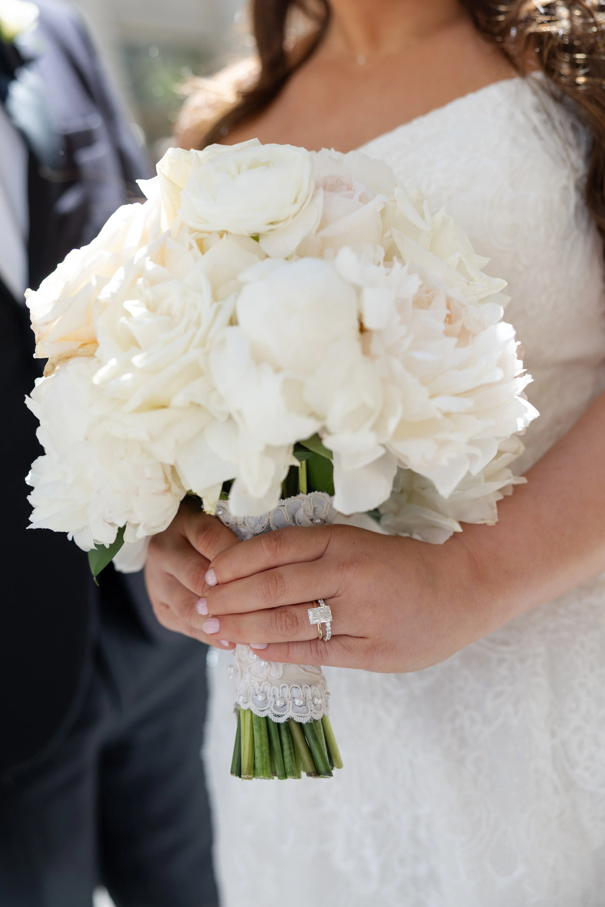 A bride holding a bouquet of white flowers, wearing a wedding dress with lace details, shows her hand with a diamond engagement ring. Part of a groom dressed in a gray suit is visible beside her.