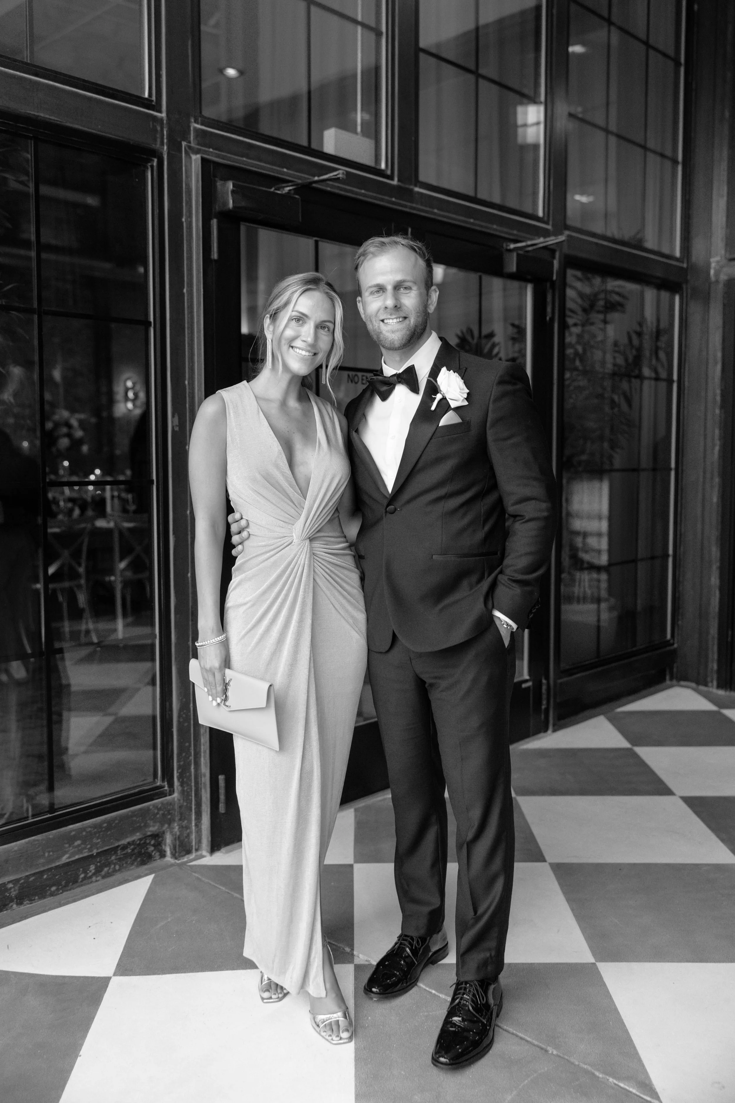 Black and white photo of a smiling couple dressed for a formal event, standing in front of a large glass door. The woman is wearing a sleeveless gown and holding a clutch, while the man is in a tuxedo with a bow tie and boutonniere.