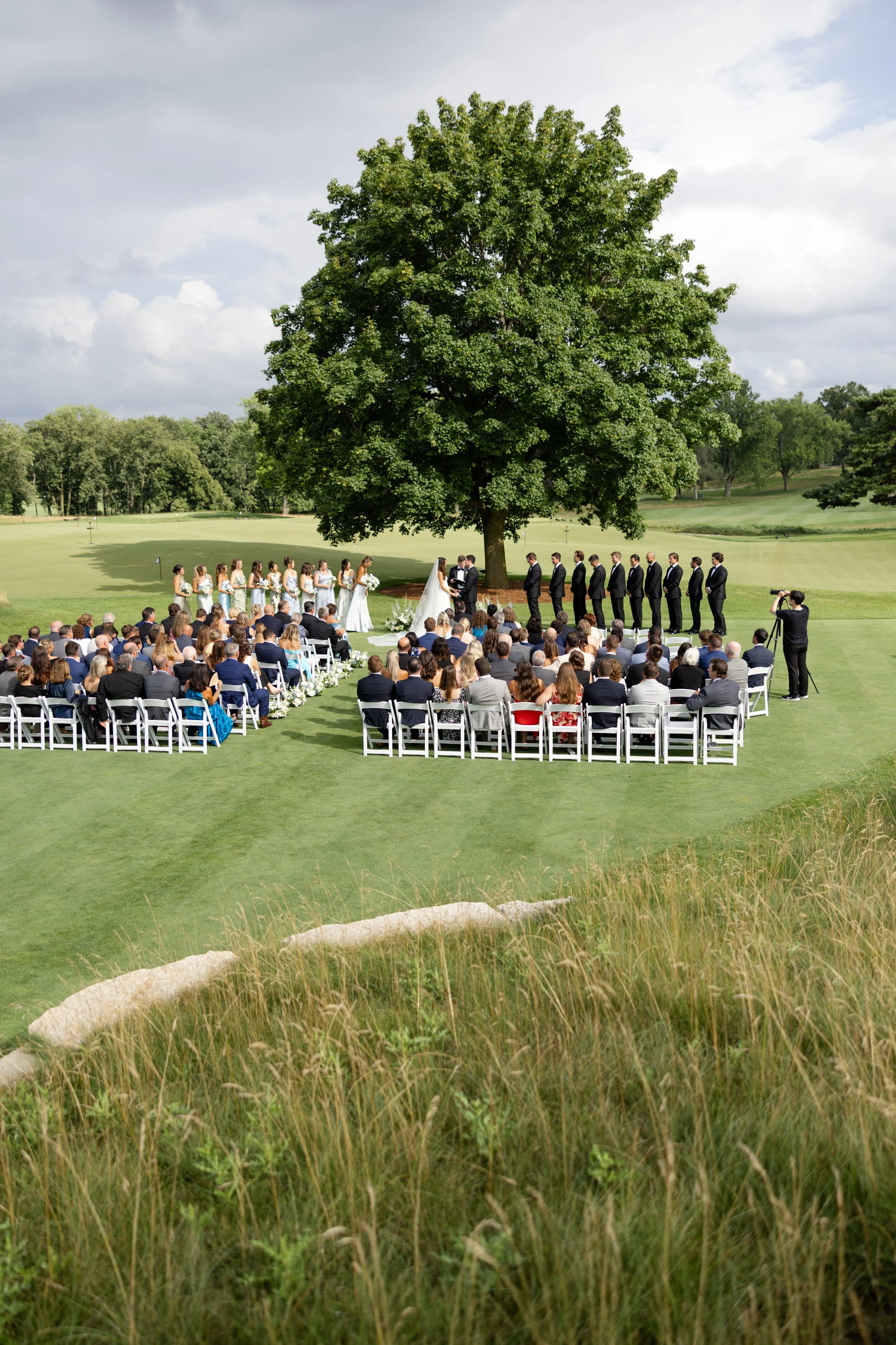 Wedding ceremony outdoors under a large tree with guests seated in white chairs, couple standing in front of officiant, photographers capturing the moment, in a lush green landscape.