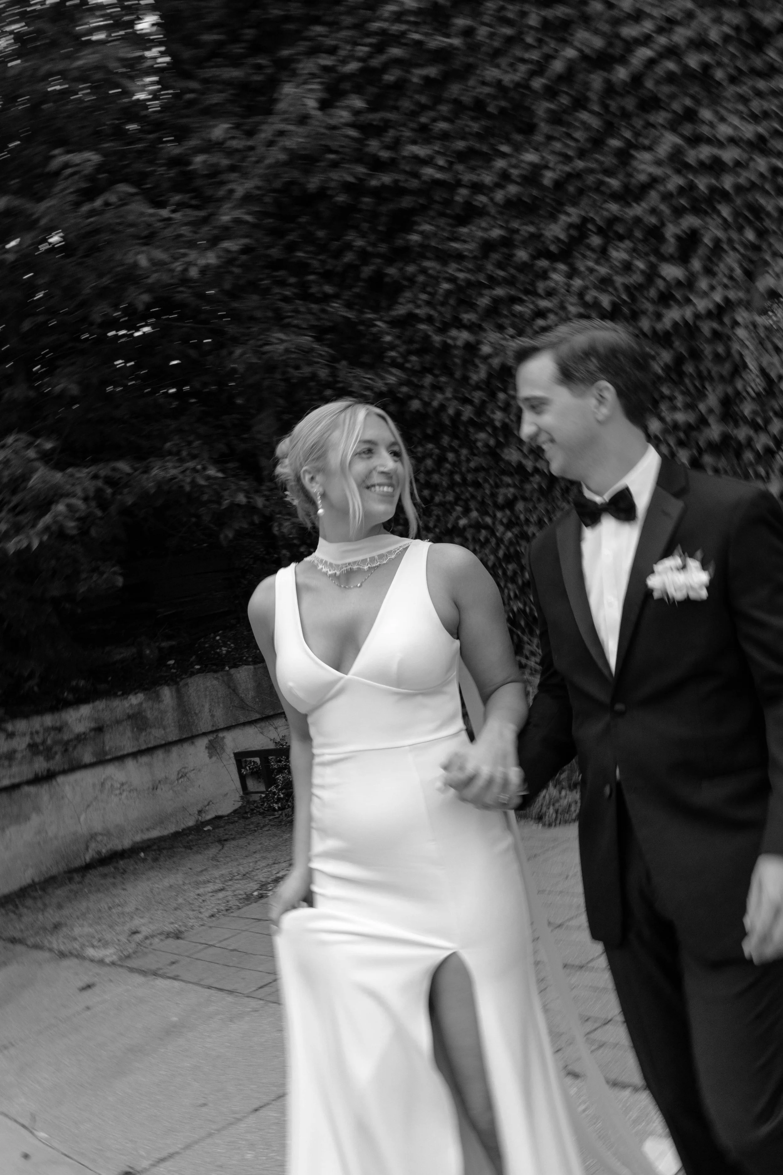A smiling couple dressed in wedding attire, holding hands and walking outdoors in front of a leafy hedge.