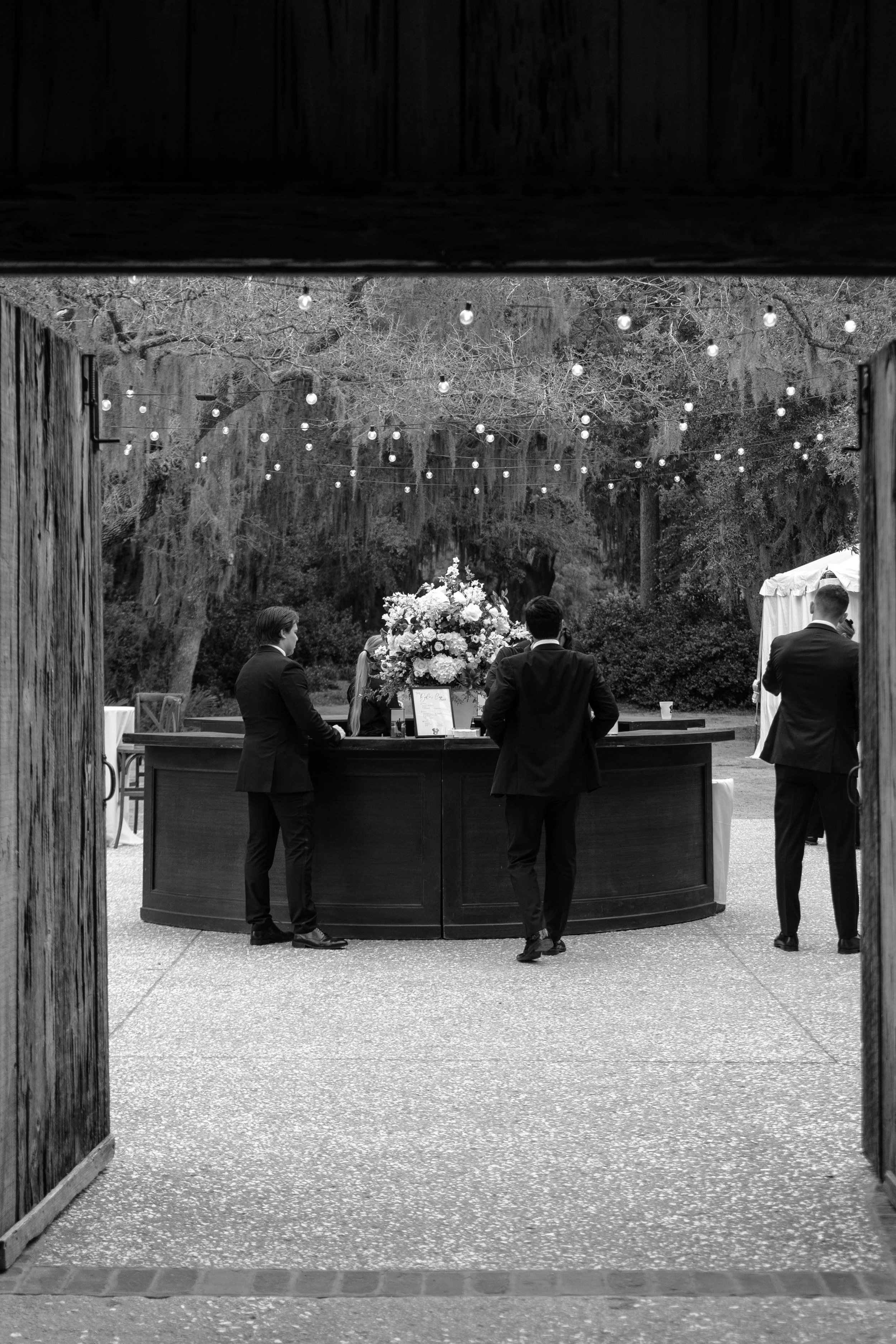 Black and white photo of a wedding reception with guests in tuxedos, a large floral centerpiece, and string lights hanging above outdoors, framed by a wooden door.