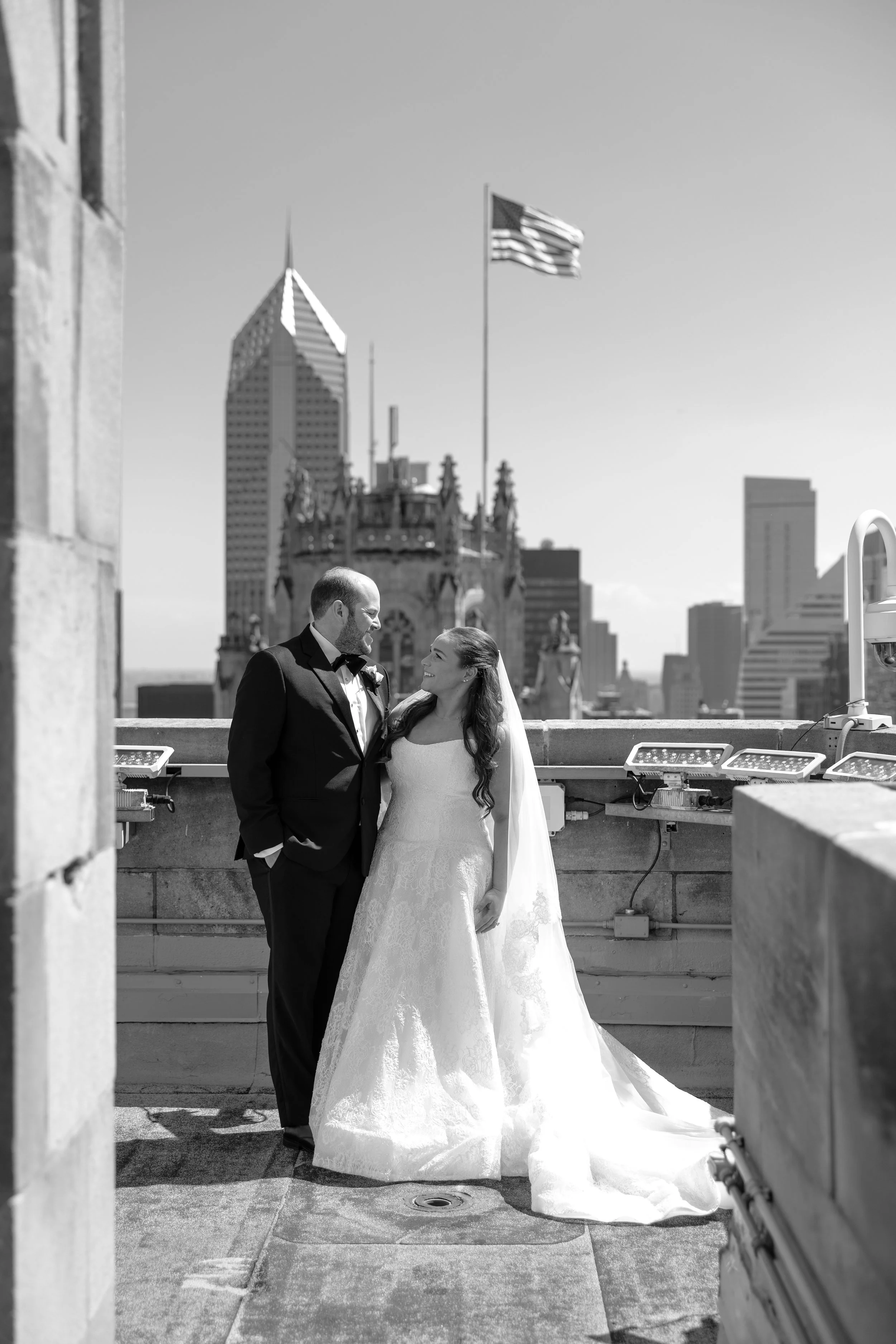 A bride and groom standing close together on a rooftop with city skyscrapers in the background, smiling at each other.