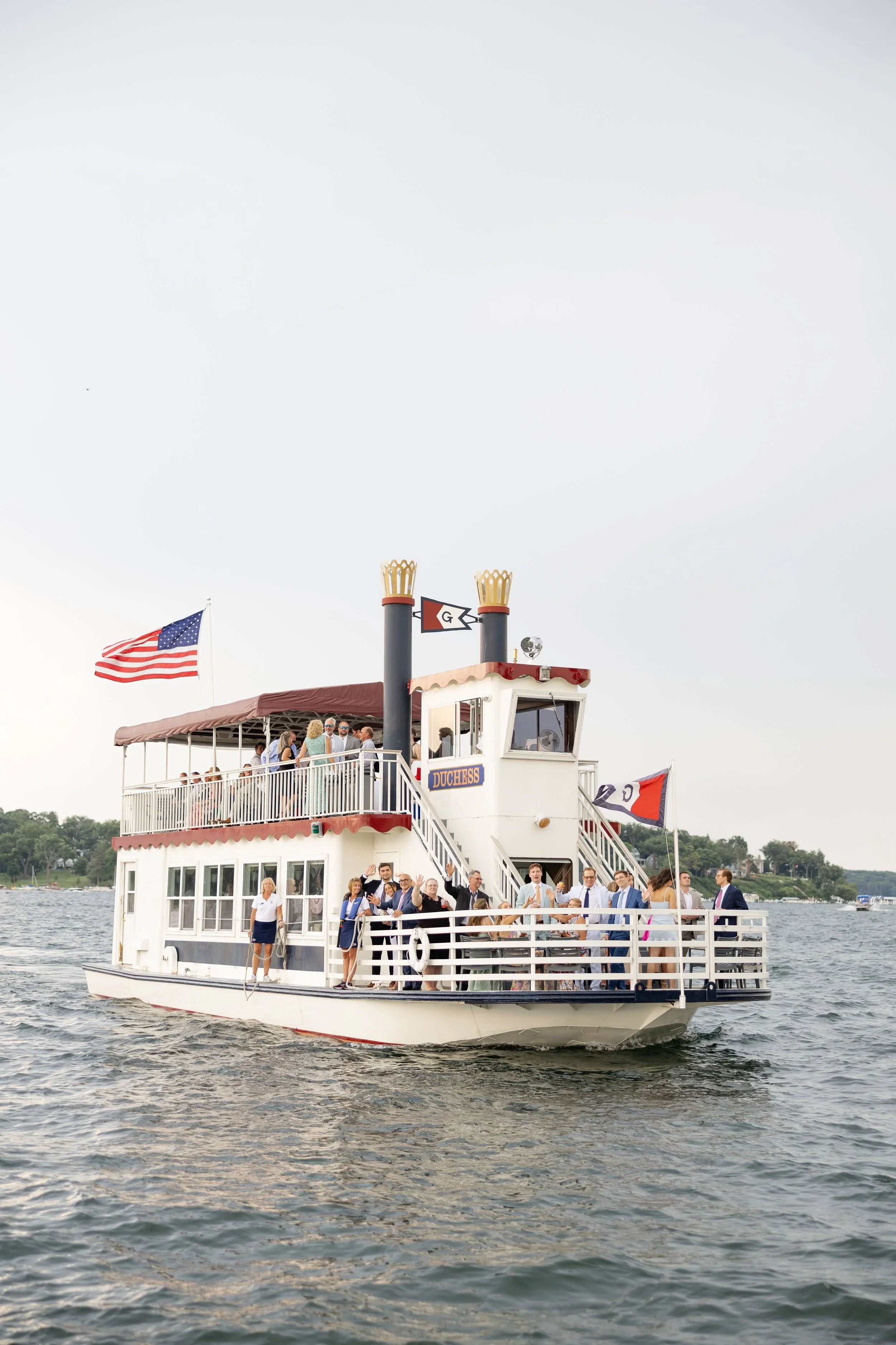 A boat on the water carrying people, with the American flag visible on the left and a flag with a black arrow and the letter G above the boat. The boat has a two-level structure with a maroon canopy, and the upper deck is crowded with people. The scene depicts a daytime outdoor gathering on a lake or river.