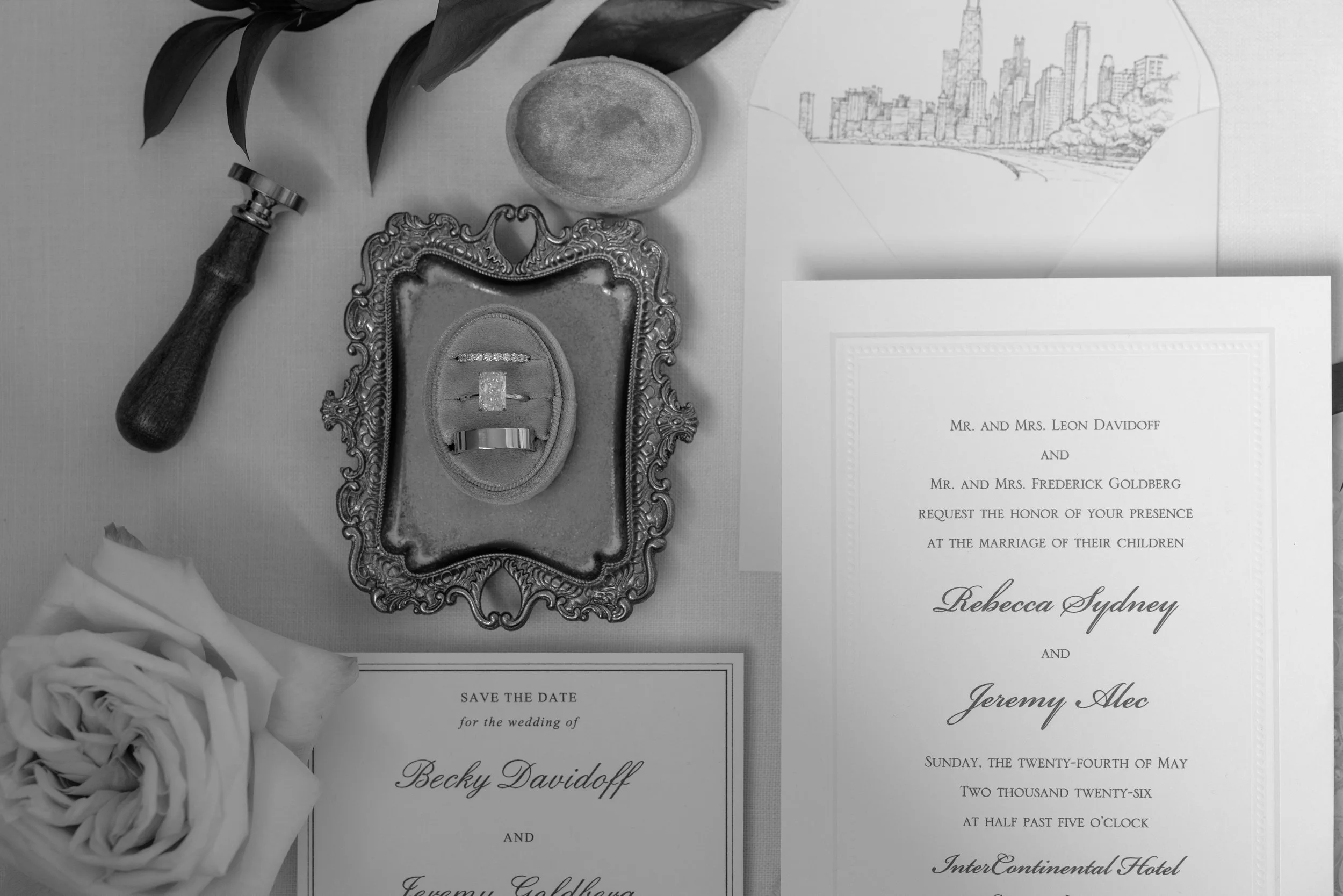 A black and white photo of wedding details including a wedding invitation, an ornate ring box with rings, a bouquet of roses, a vintage stamp, and a sketch of a city skyline.