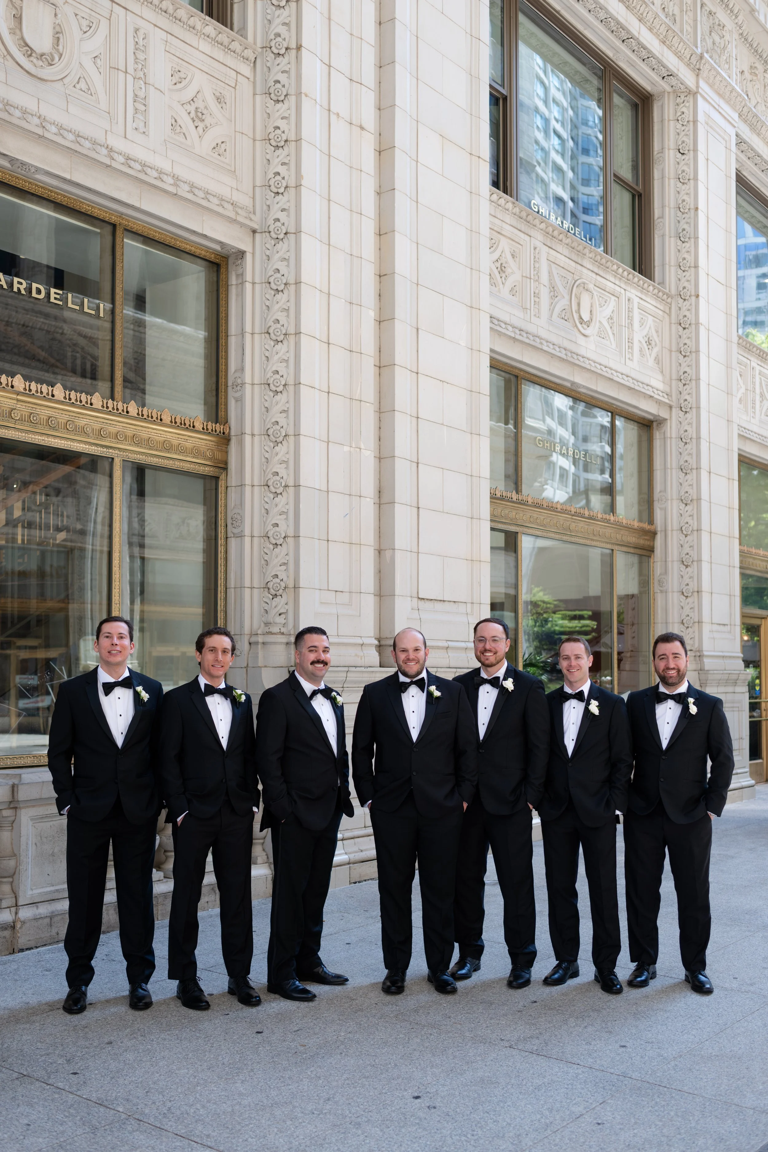 Group of seven men dressed in black tuxedos with white shirts and black bow ties, standing outdoors in front of a building with ornate stone architecture and large glass windows. The men are smiling and appear to be at a formal event.