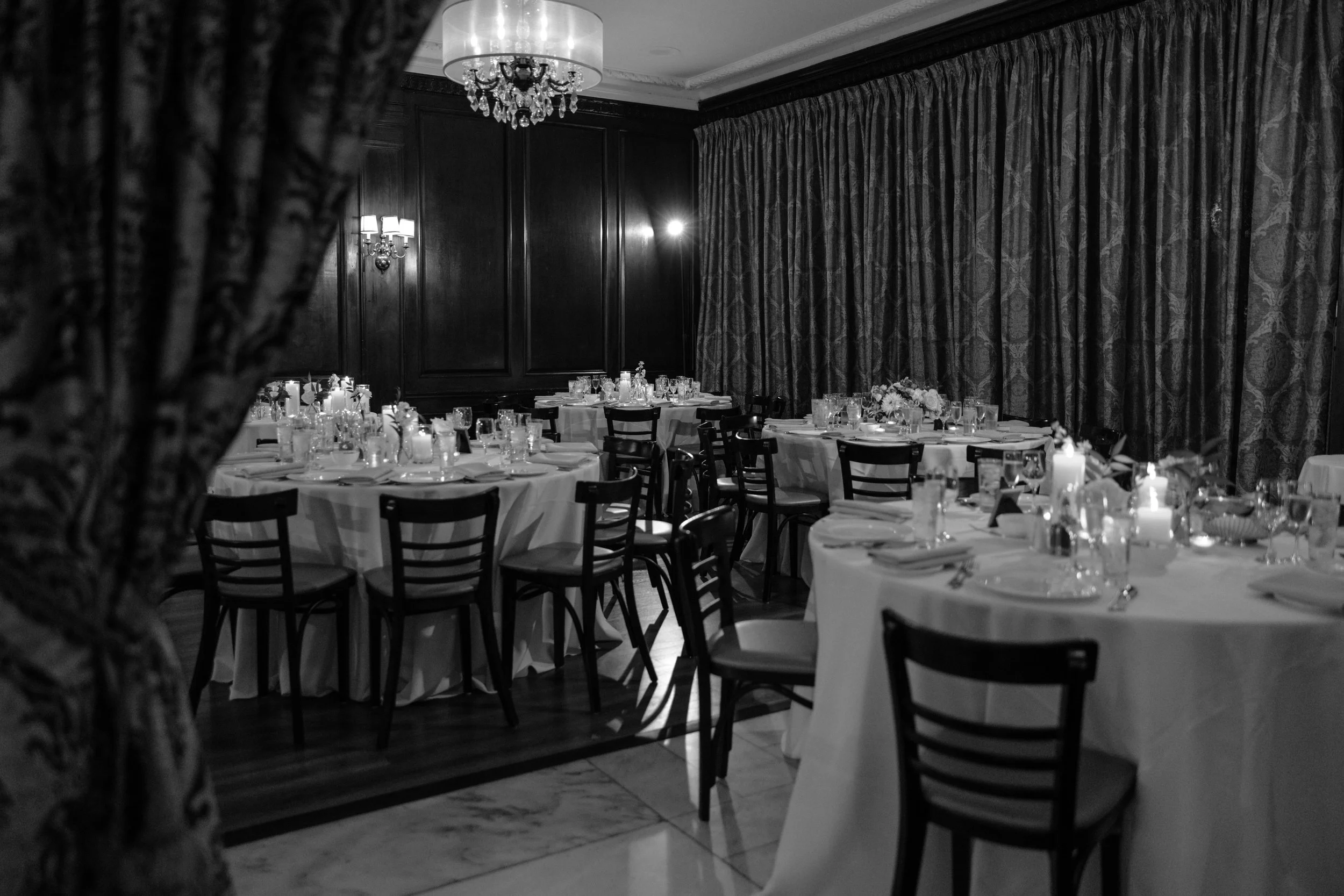 Black and white photo of a decorated banquet room with round tables set for an event, surrounded by chairs, with elegant curtains and lighting fixtures on dark wood-paneled walls.