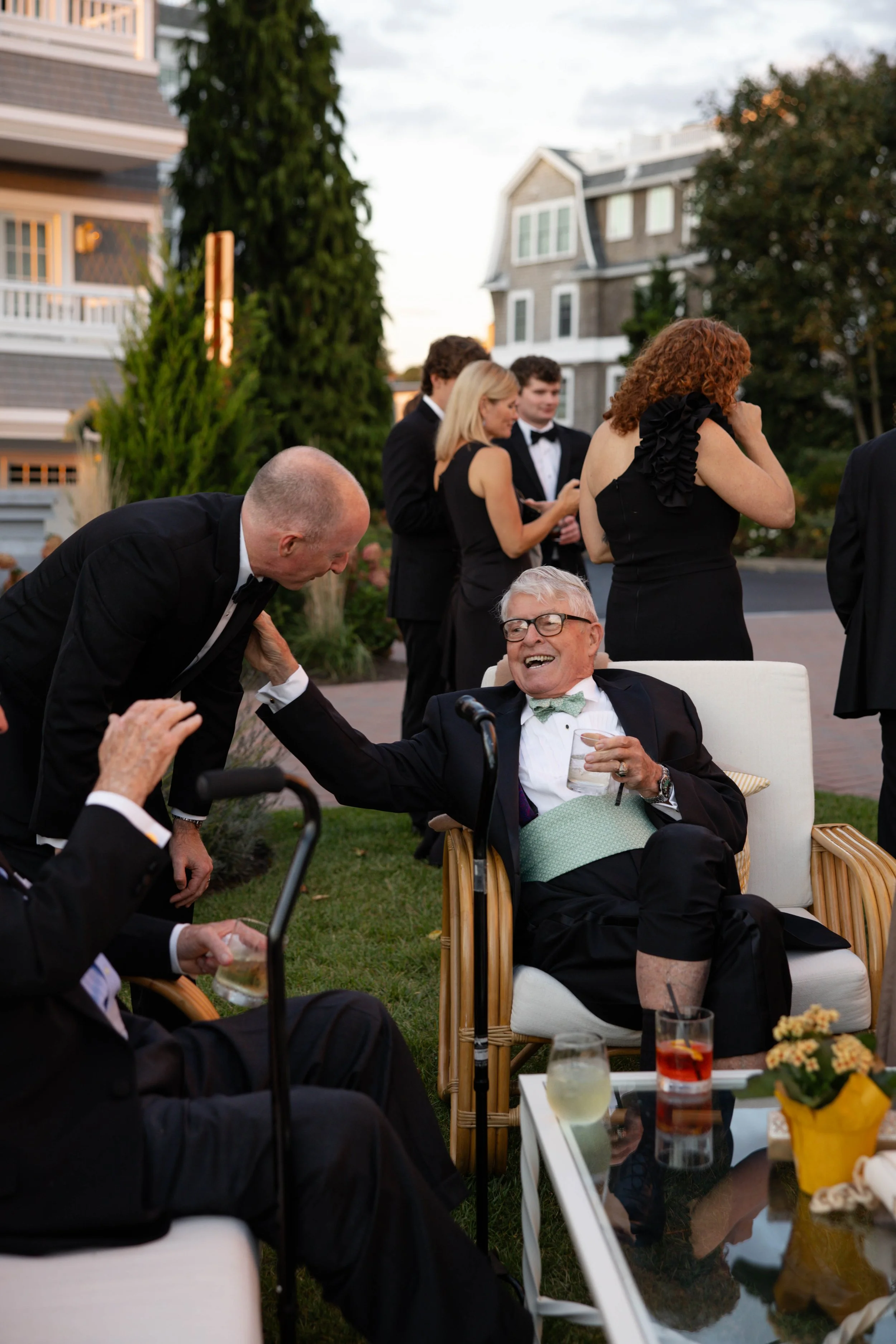 A group of people, dressed in formal evening wear, socializing outdoors in a backyard during the evening. Some are sitting on chairs, while others stand and chat, with a house and trees in the background.