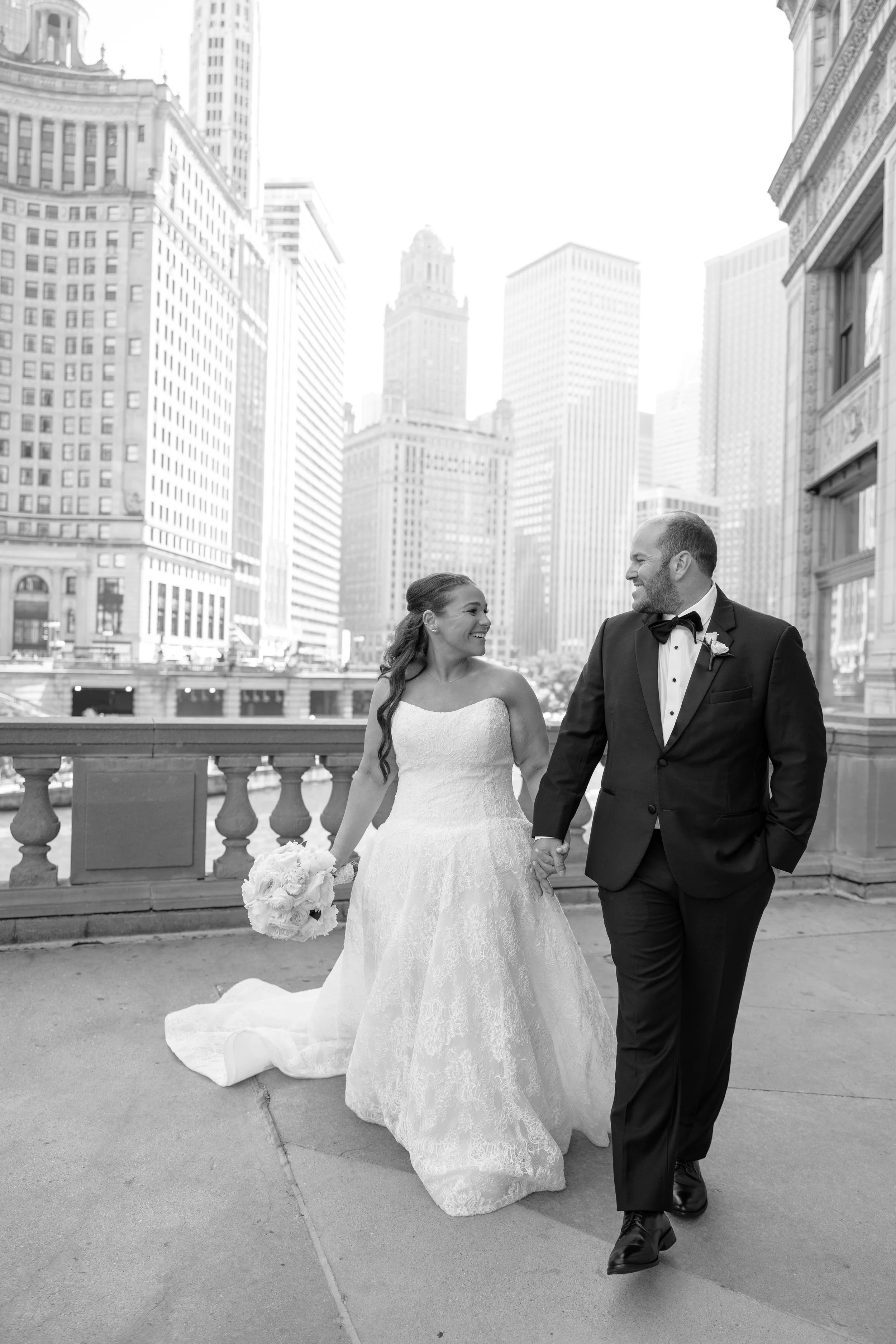 A bride and groom walking outside in an urban cityscape. The bride is wearing a lace wedding dress and holding a bouquet of flowers, and the groom is dressed in a tuxedo. They are holding hands and smiling at each other with skyscrapers in the backgr