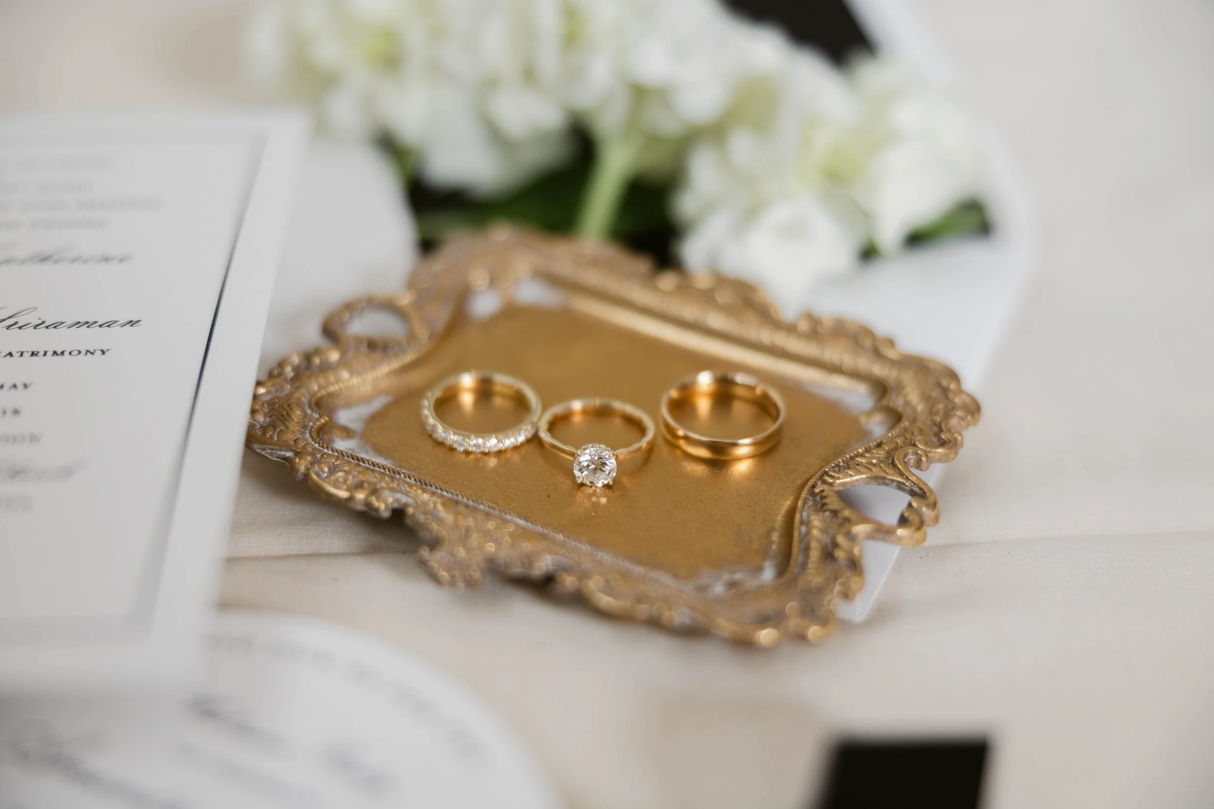 Close-up of gold wedding rings and an engagement ring with a large diamond, placed on an ornate gold tray. In the background, white flowers and a part of a wedding invitation are visible.
