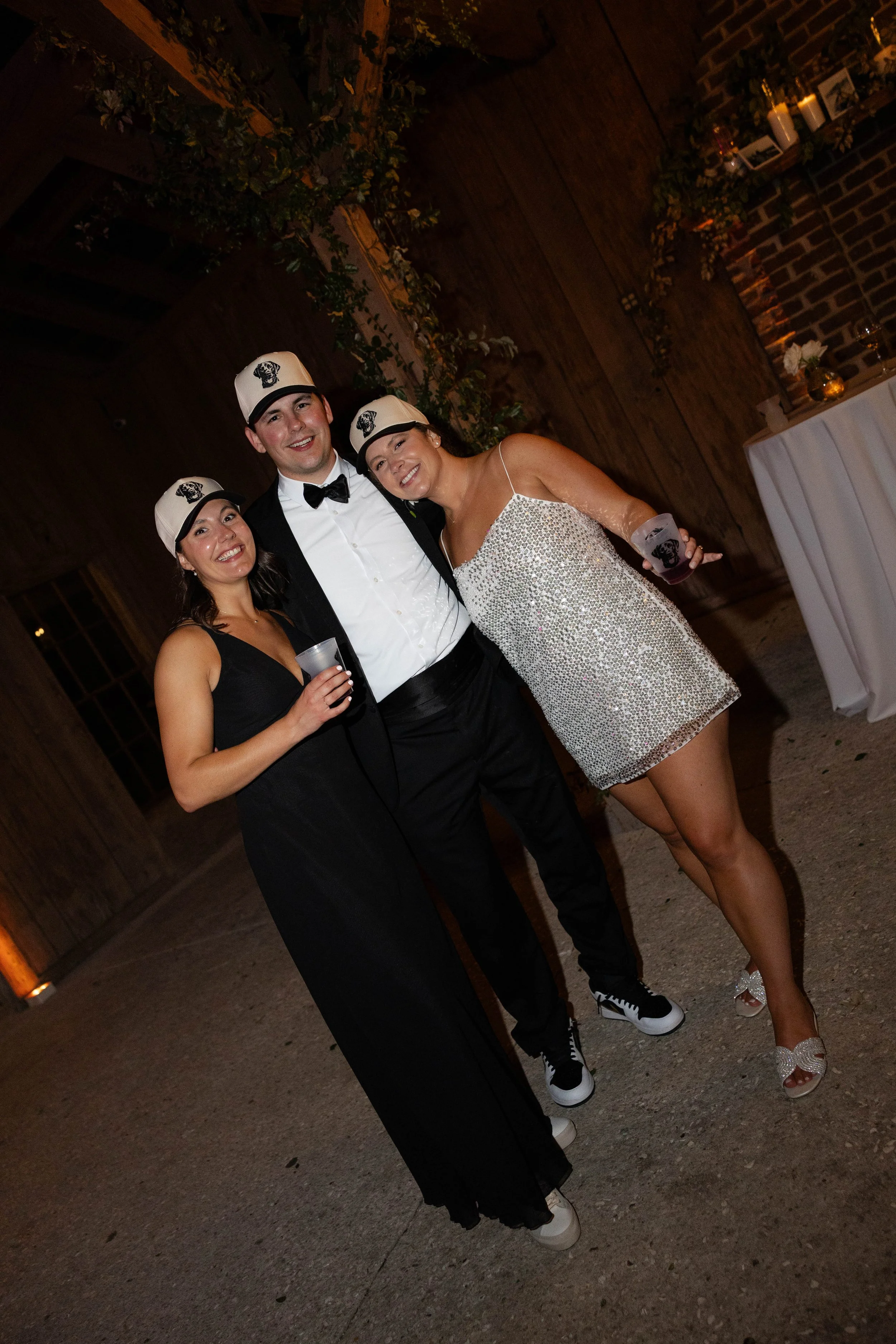 Three people smiling and posing for a photo at a party. They are wearing matching white caps with black logos. The person on the left wears a black dress, the person in the middle wears a tuxedo with a bow tie, and the person on the right wears a spa