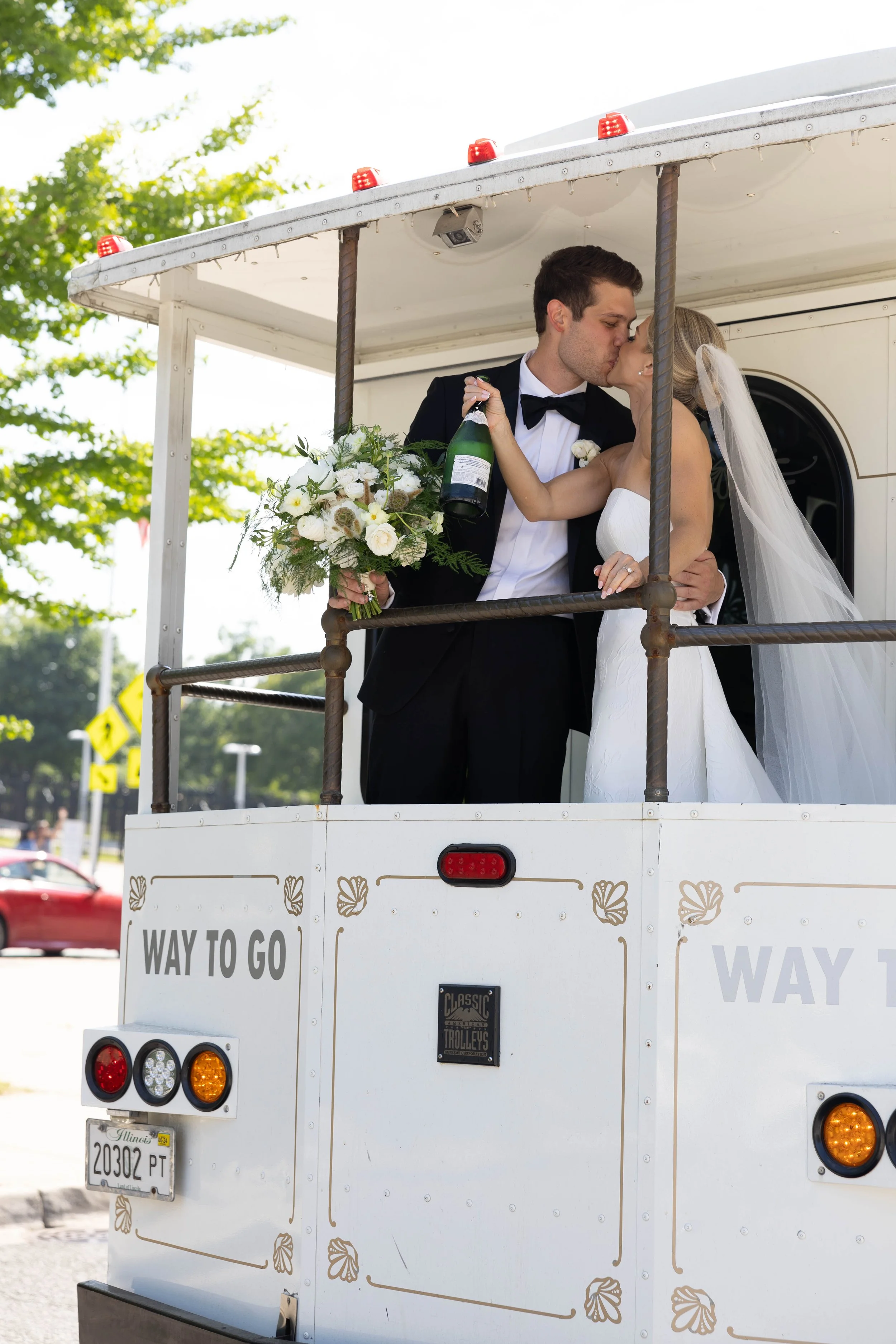 A bride and groom kissing on a white trolley decorated for a wedding, with the bride holding a bouquet of white and green flowers and the groom holding a champagne bottle, under a bright, sunny sky.