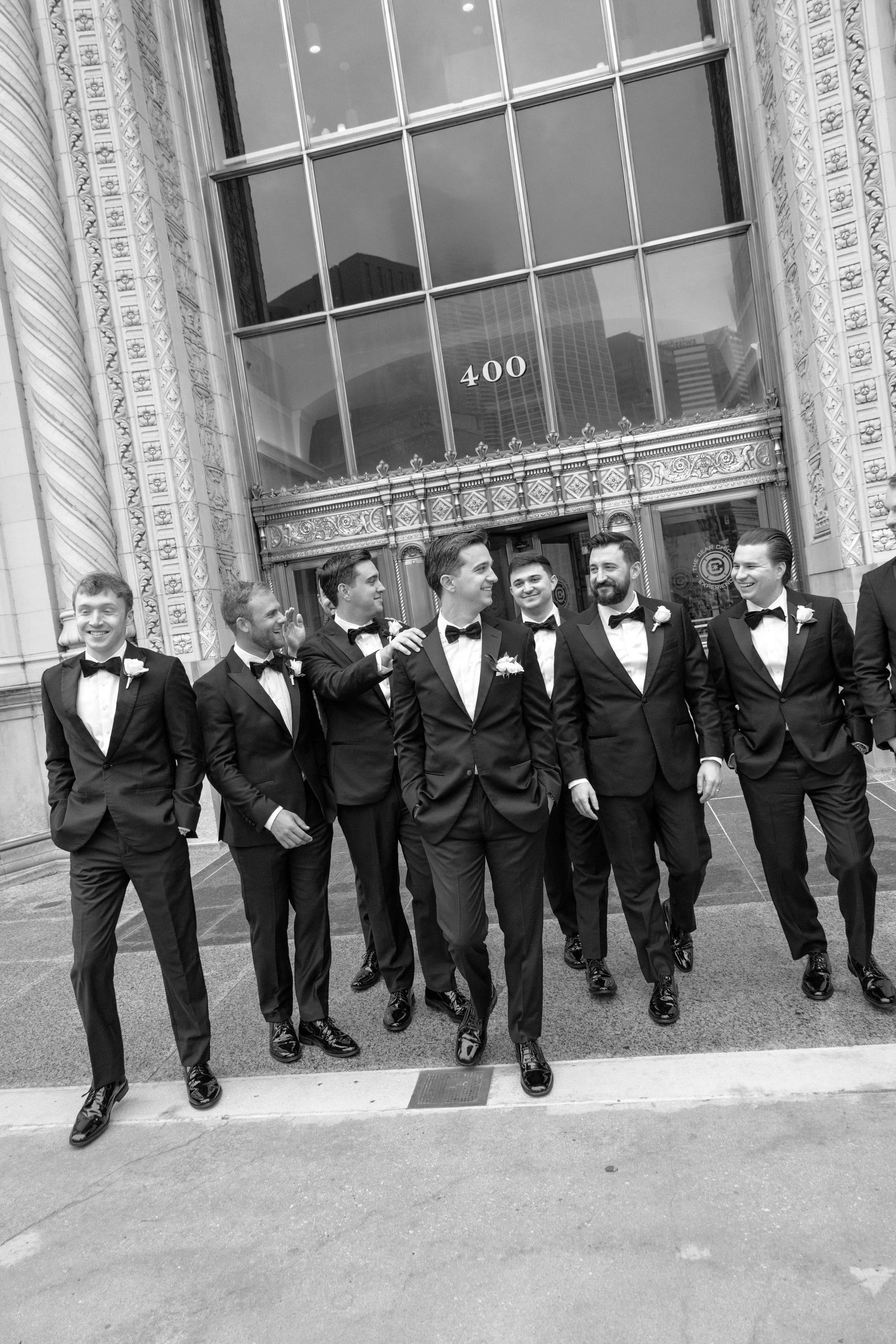A group of men in tuxedos and bow ties walking in front of an ornate building with large glass windows and the number 400 displayed. The men are smiling and appear to be in formal attire, possibly for a wedding or formal event.