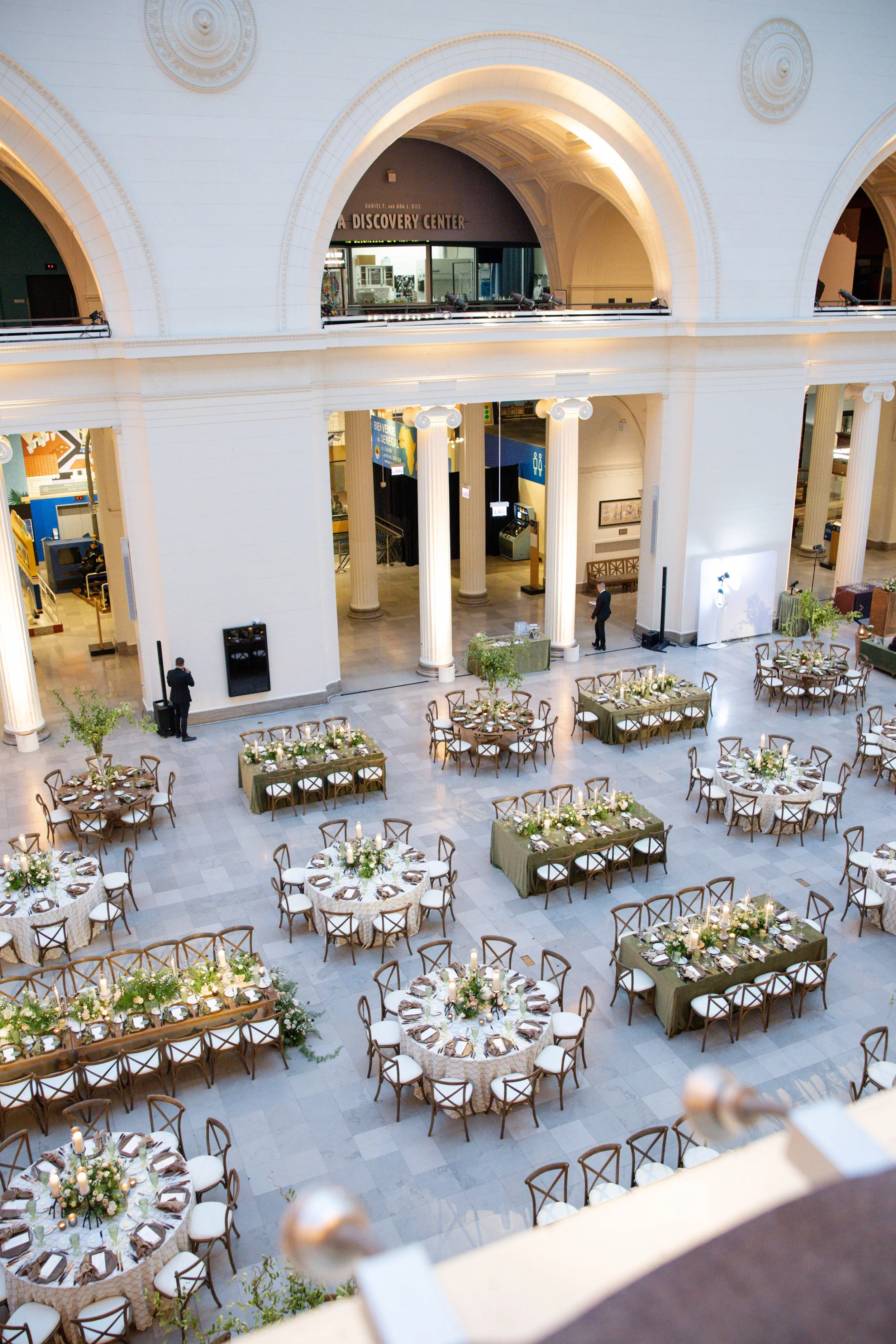 Large indoor space with high ceilings, columns, and arches, set up for a formal event with round and rectangular tables decorated with flowers and candles, and elegant chairs, possibly a banquet or wedding reception.