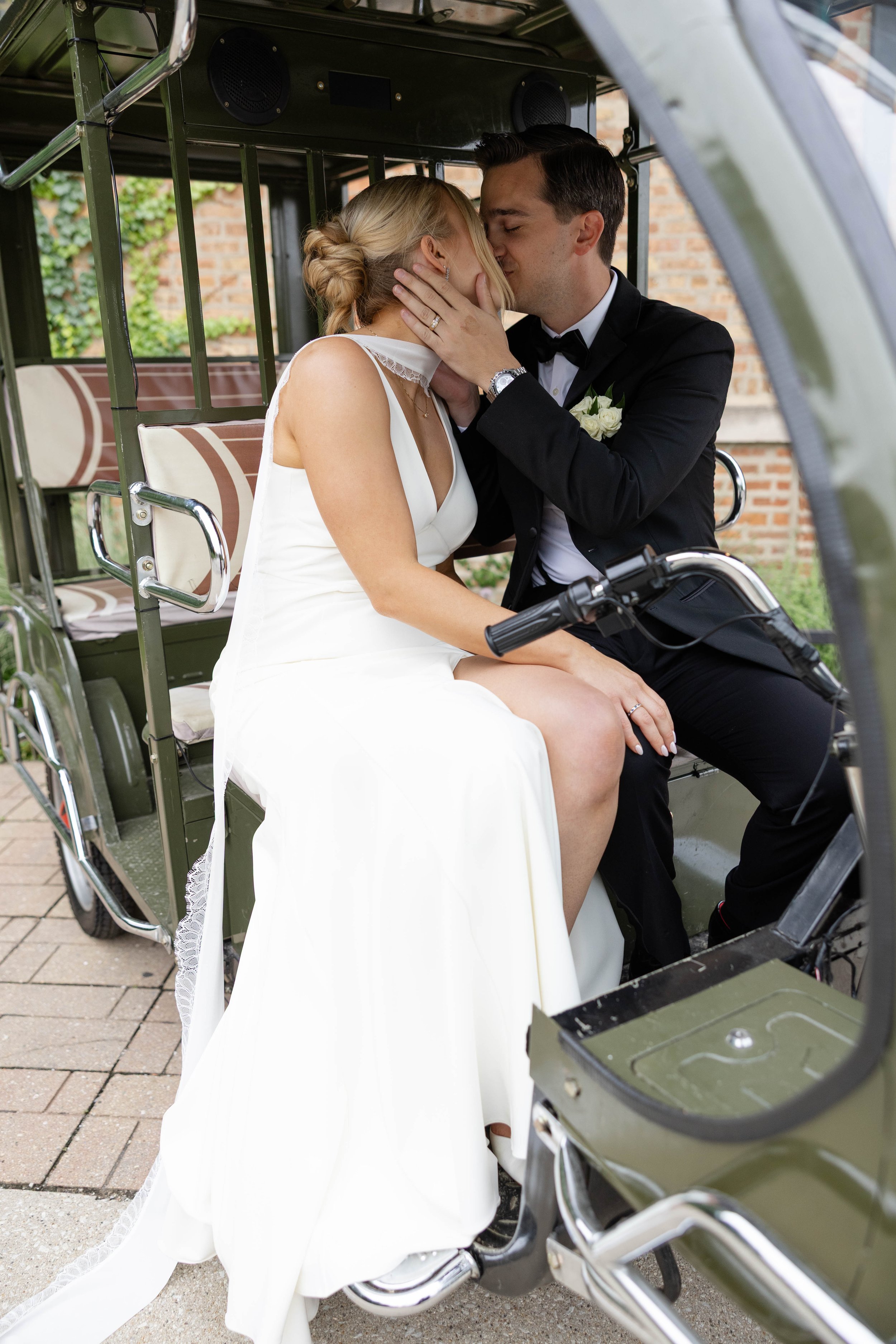 A bride and groom sharing a kiss inside a motorized rickshaw or tuk-tuk. The bride is wearing a white wedding dress, and the groom is dressed in a black tuxedo with a boutonnière. The scene is set outdoors near a brick building.