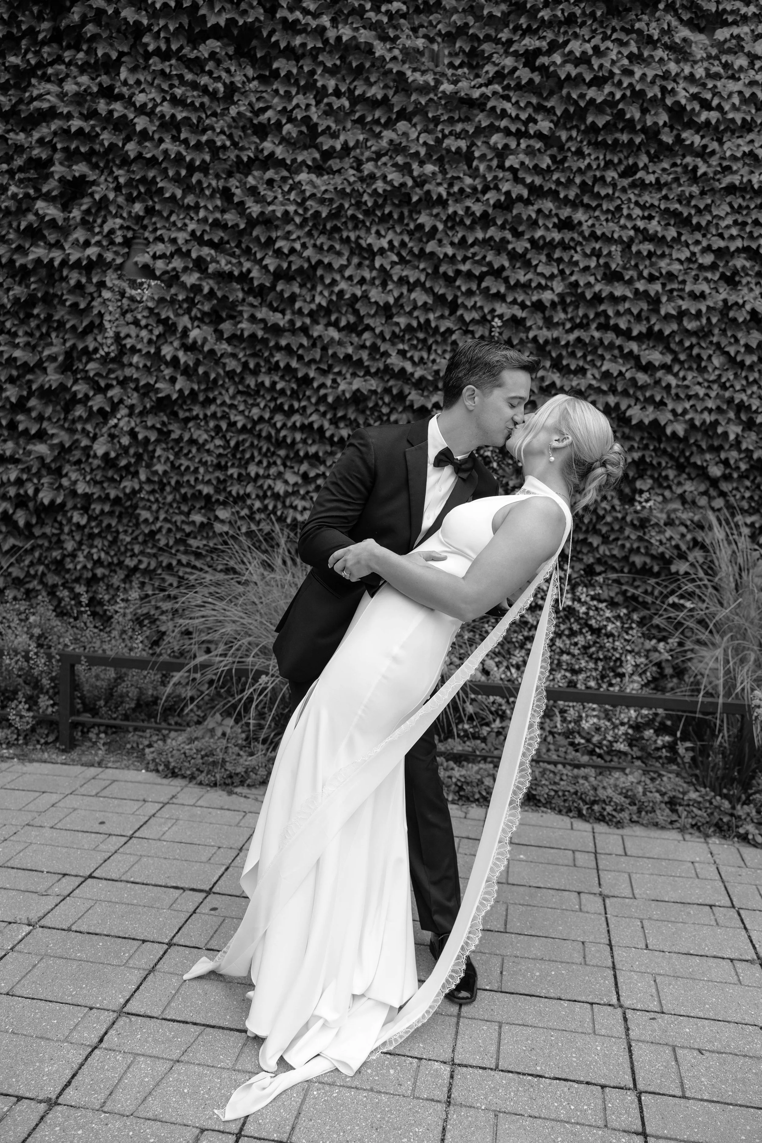 A black-and-white photo of a bride and groom kissing outside against a hedge wall. The groom, in a tuxedo with a bow tie, is leaning down to kiss the bride, who is in a sleeveless wedding gown. The bride is holding a ribboned bouquet, and the groom i