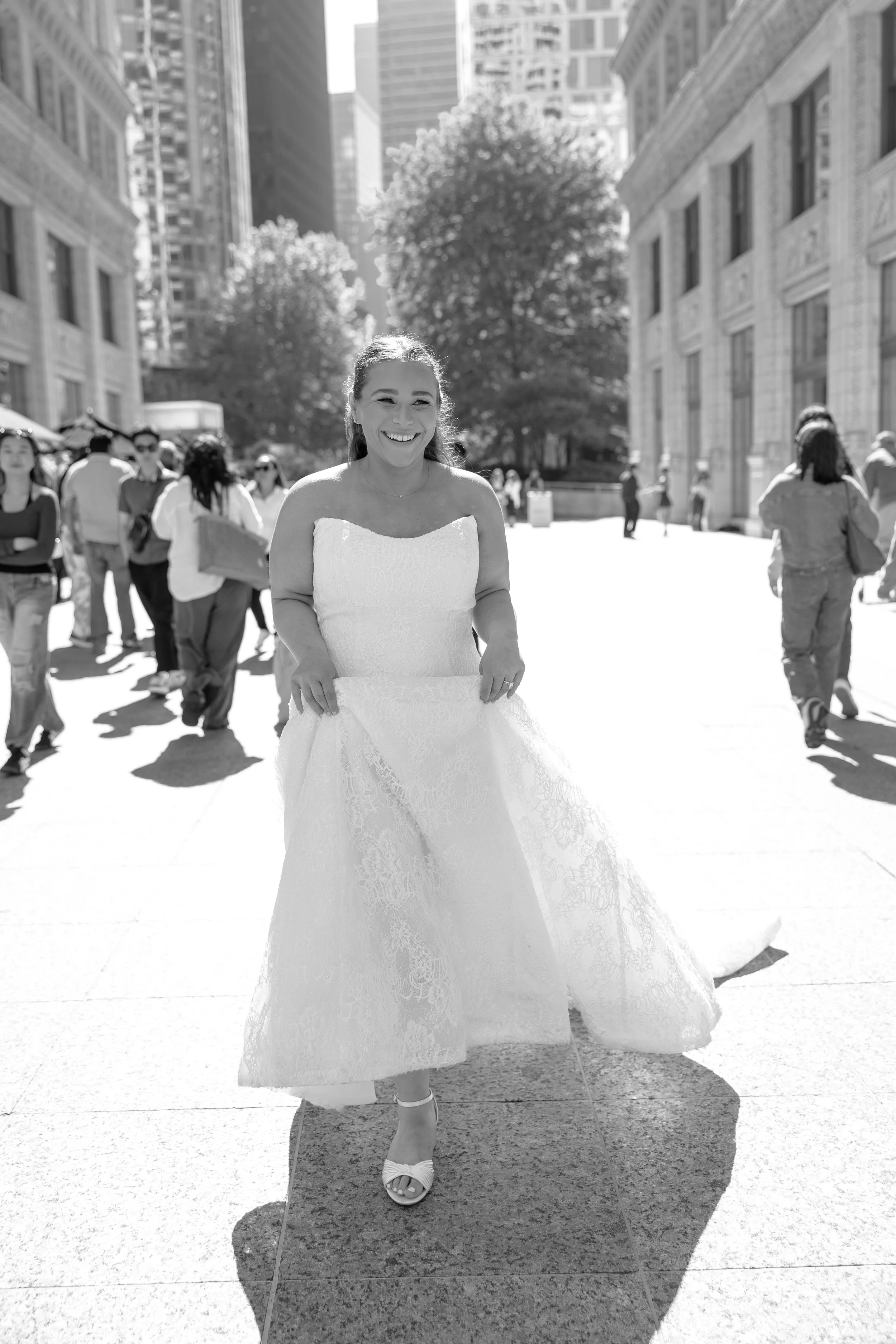 A woman in a wedding dress smiling walking on a city sidewalk with tall buildings and trees in the background, surrounded by other pedestrians.