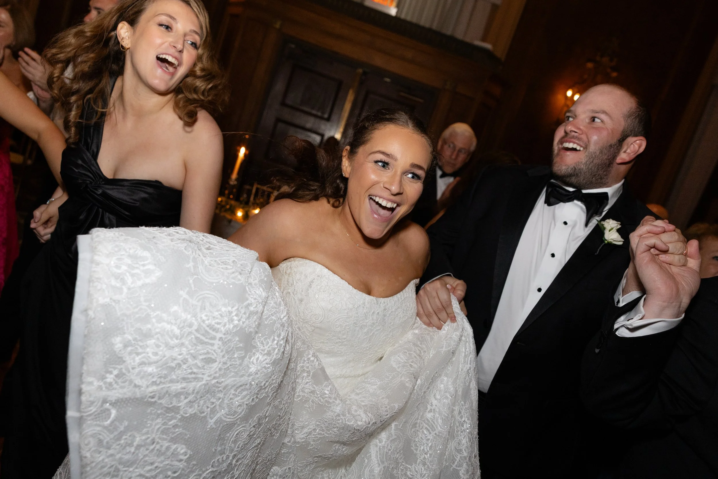 A bride and groom dancing with two smiling women at a wedding reception, with warm wooden walls and dim lighting in the background.