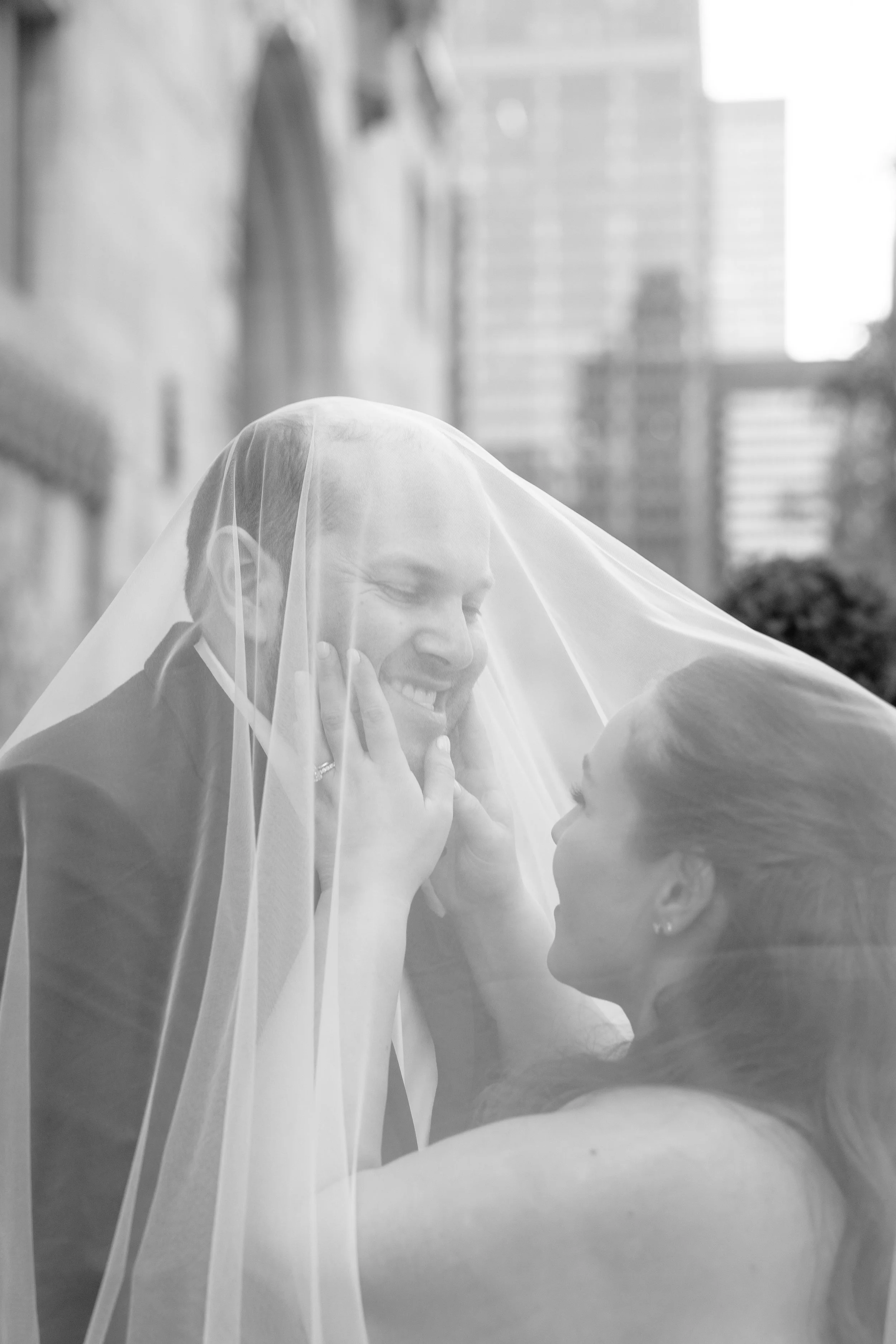 A woman with a veil talking to a young girl, both smiling, outdoors with tall buildings in the background.