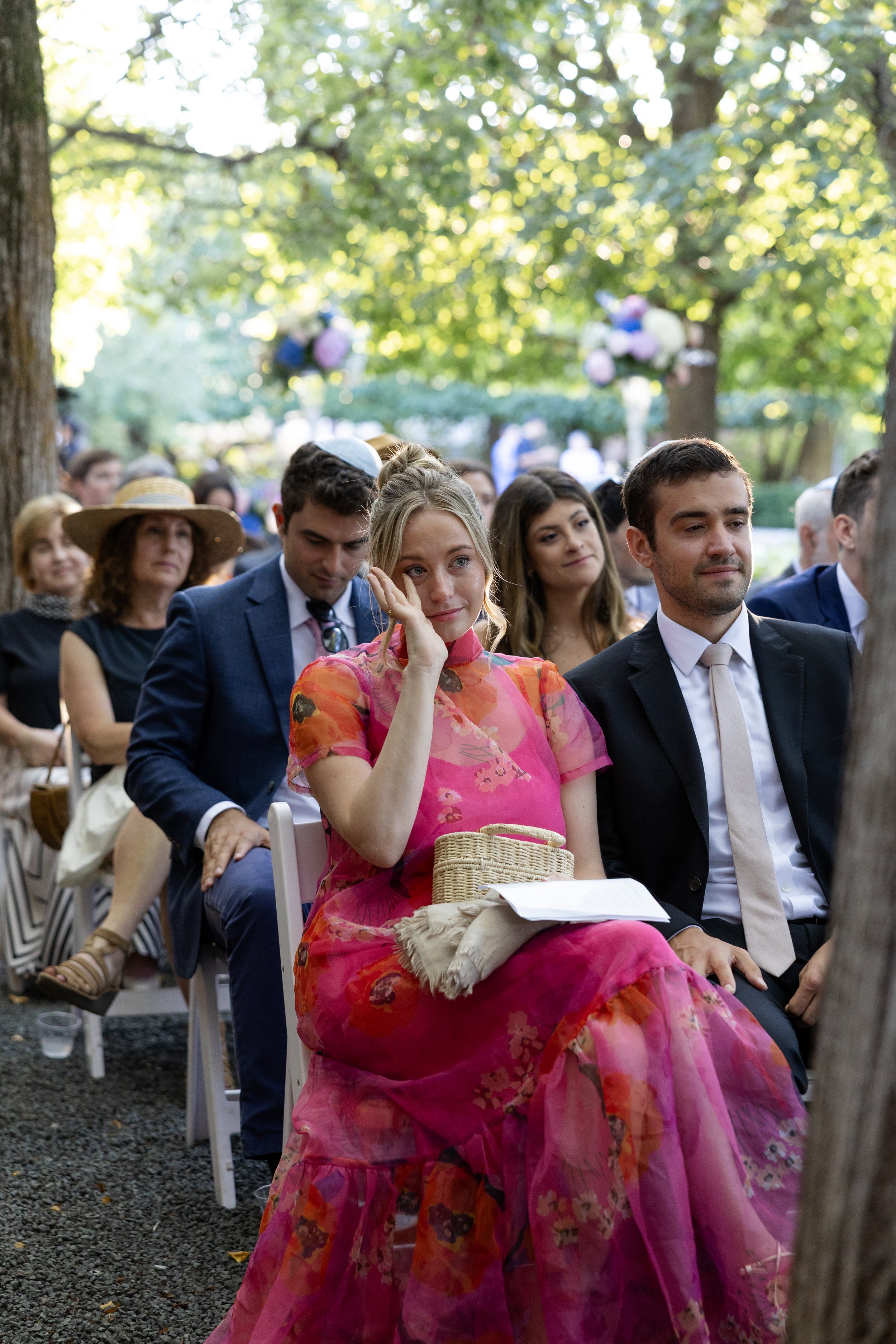 A group of people dressed in formal and semi-formal attire sitting outdoors at a gathering or event in a park with trees, sunlight, and hanging balloons in the background.