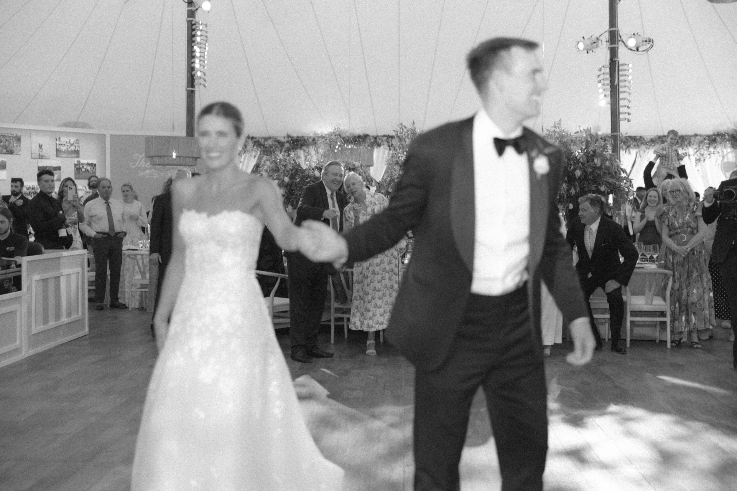 Black and white photo of a bride and groom dancing at their wedding reception, with guests in the background.
