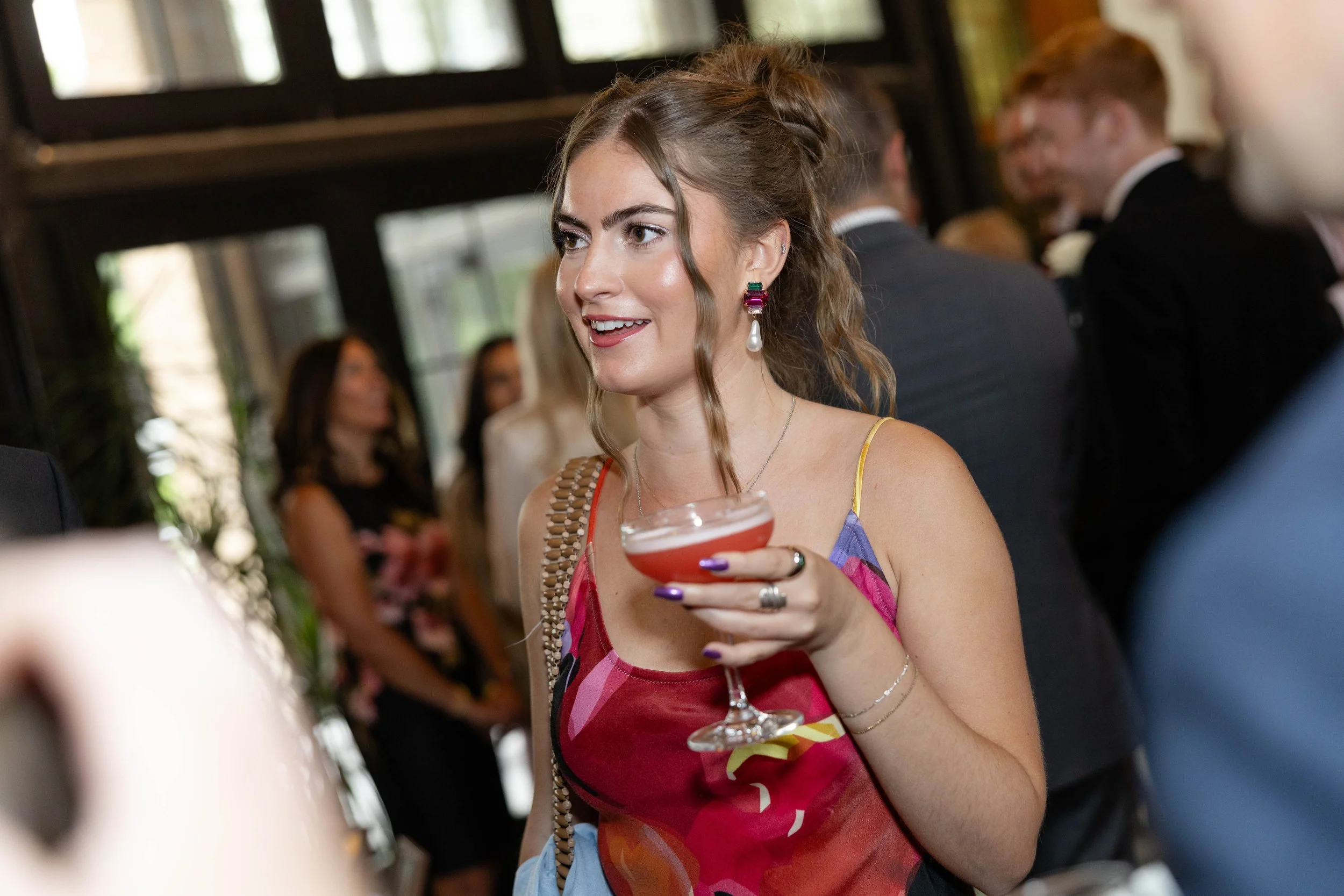 A young woman with brown wavy hair, wearing colorful earrings and a multicolored dress, holding a cocktail glass at a social gathering.