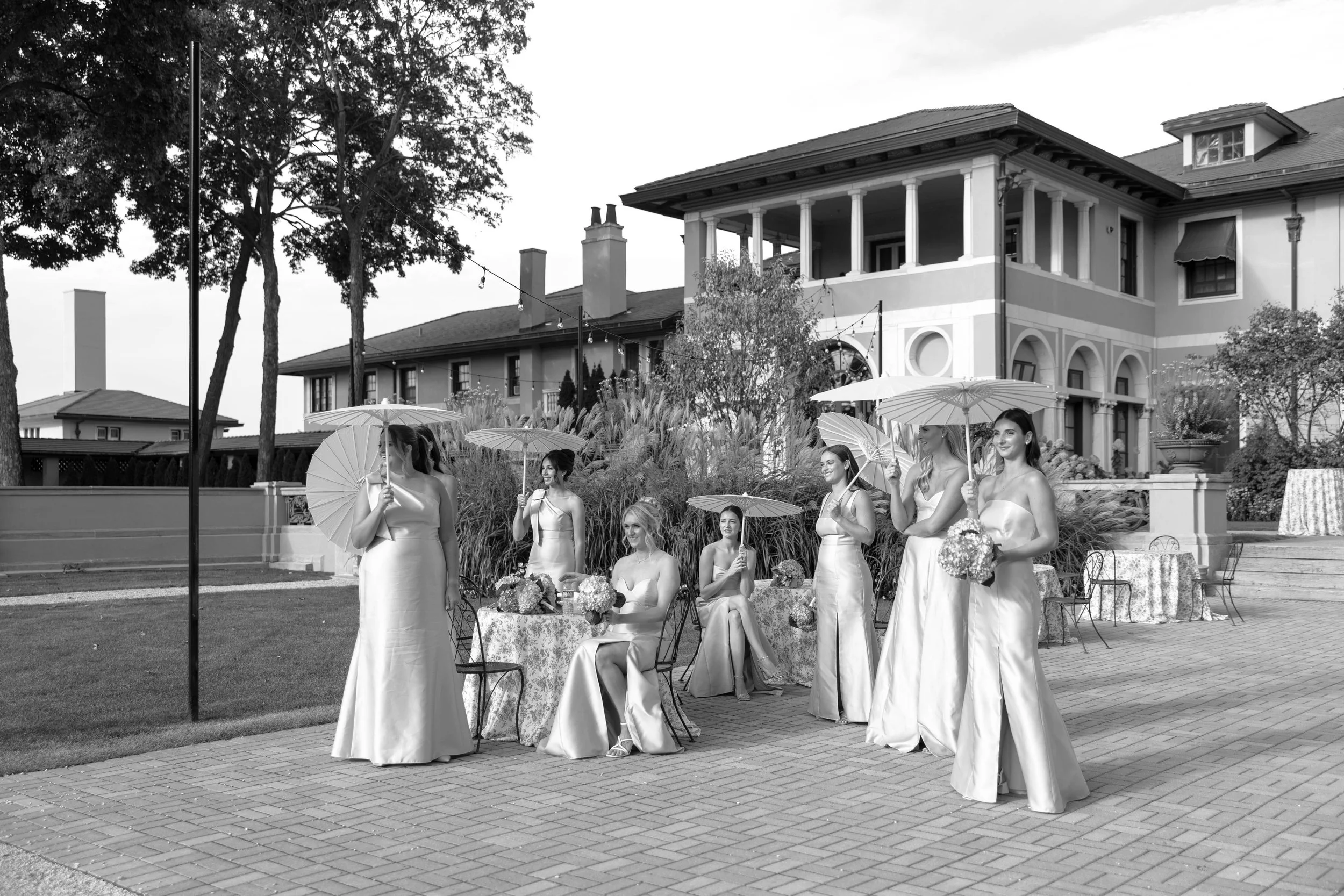 A black-and-white photo of a wedding scene outdoors. Bridesmaids dressed in long dresses holding parasols, standing around a table with flowers. A large house or mansion is visible in the background, with trees and string lights. The scene appears to be setup for a celebration or ceremony.