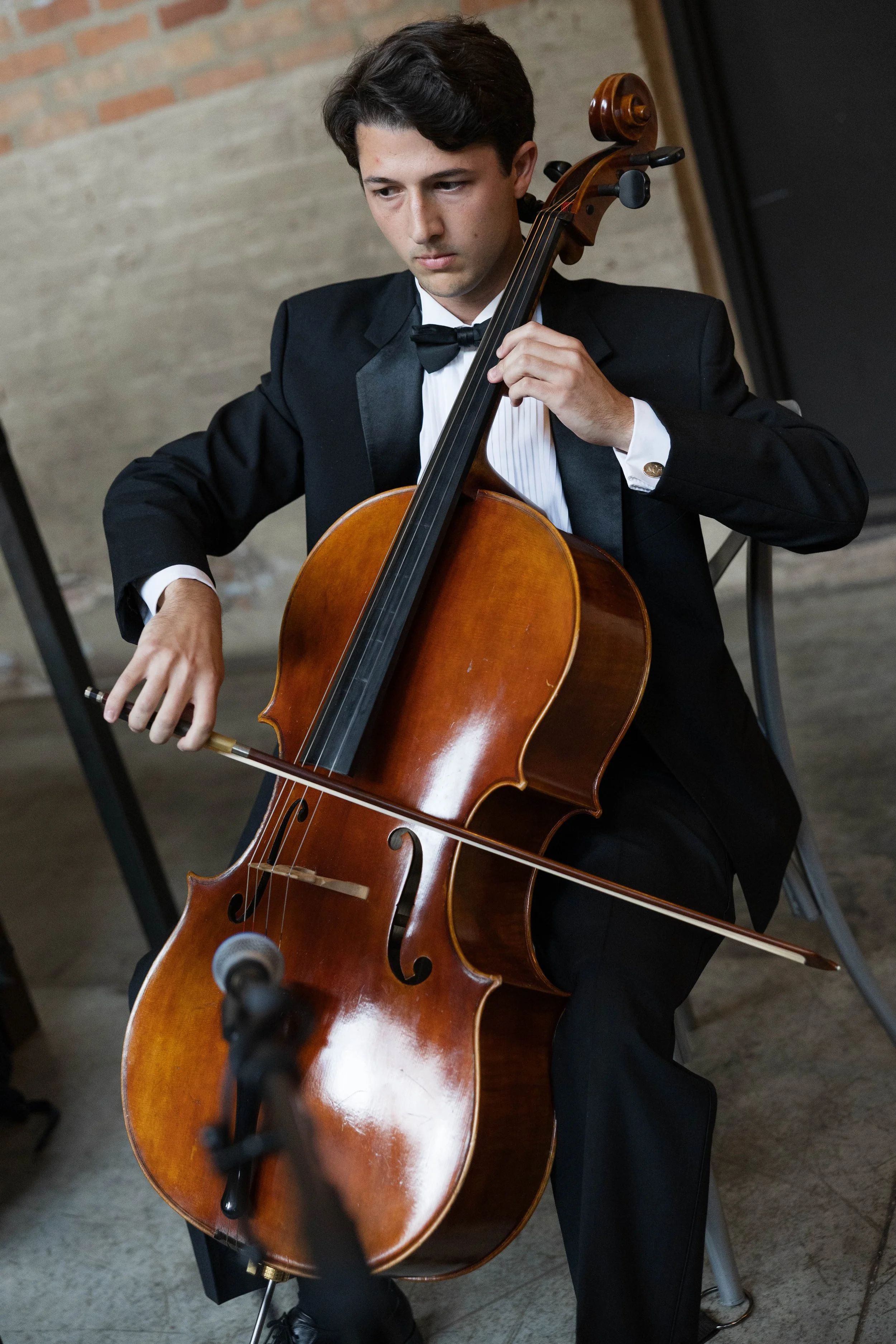 A young man in formal attire, wearing a black tuxedo with a bow tie, is playing a cello during a performance or rehearsal. He is focused on his music in an indoor setting with brick and concrete walls.