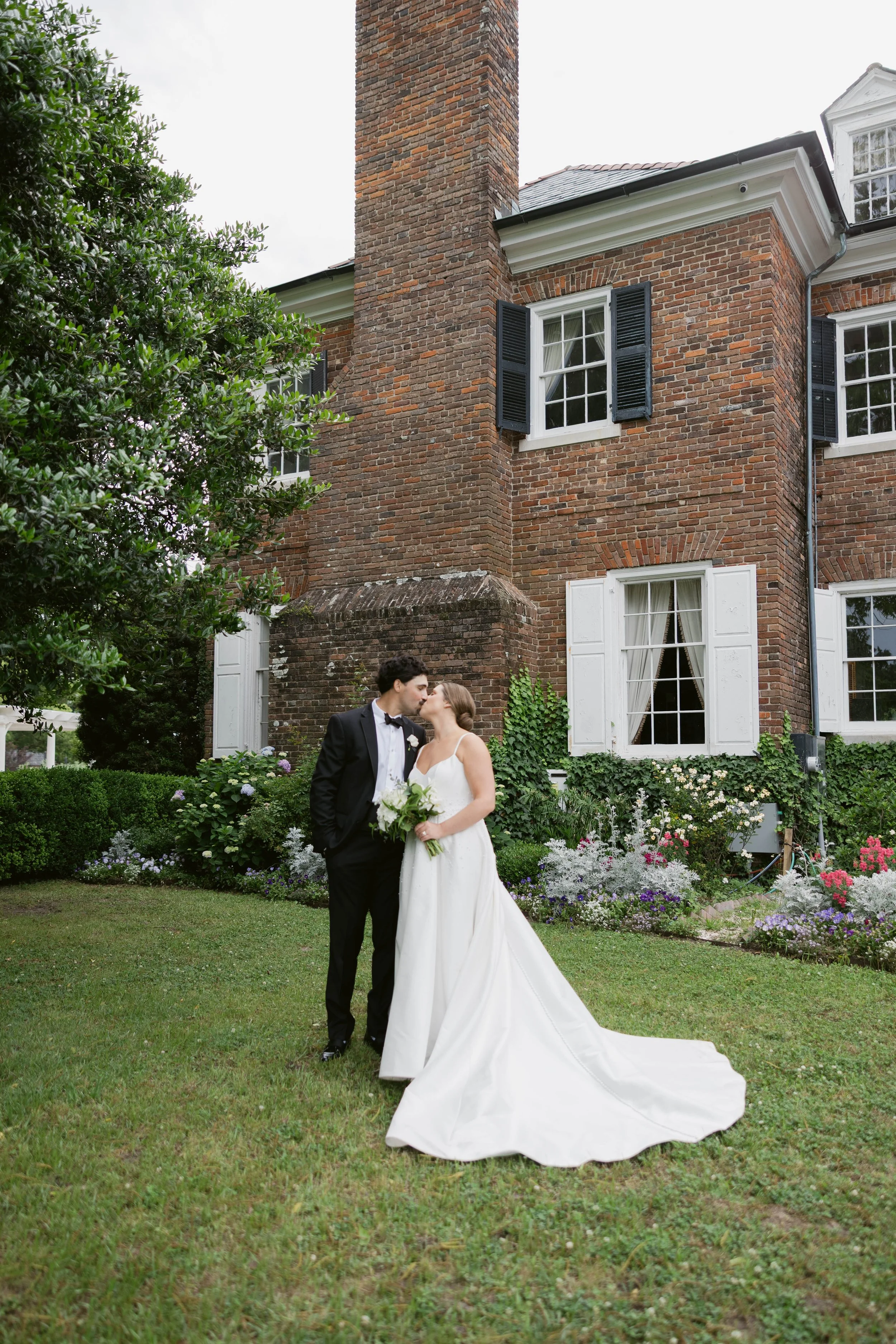 A bride and groom share a kiss outdoors in front of a brick house with white-framed windows and black shutters, surrounded by greenery and colorful flowers.