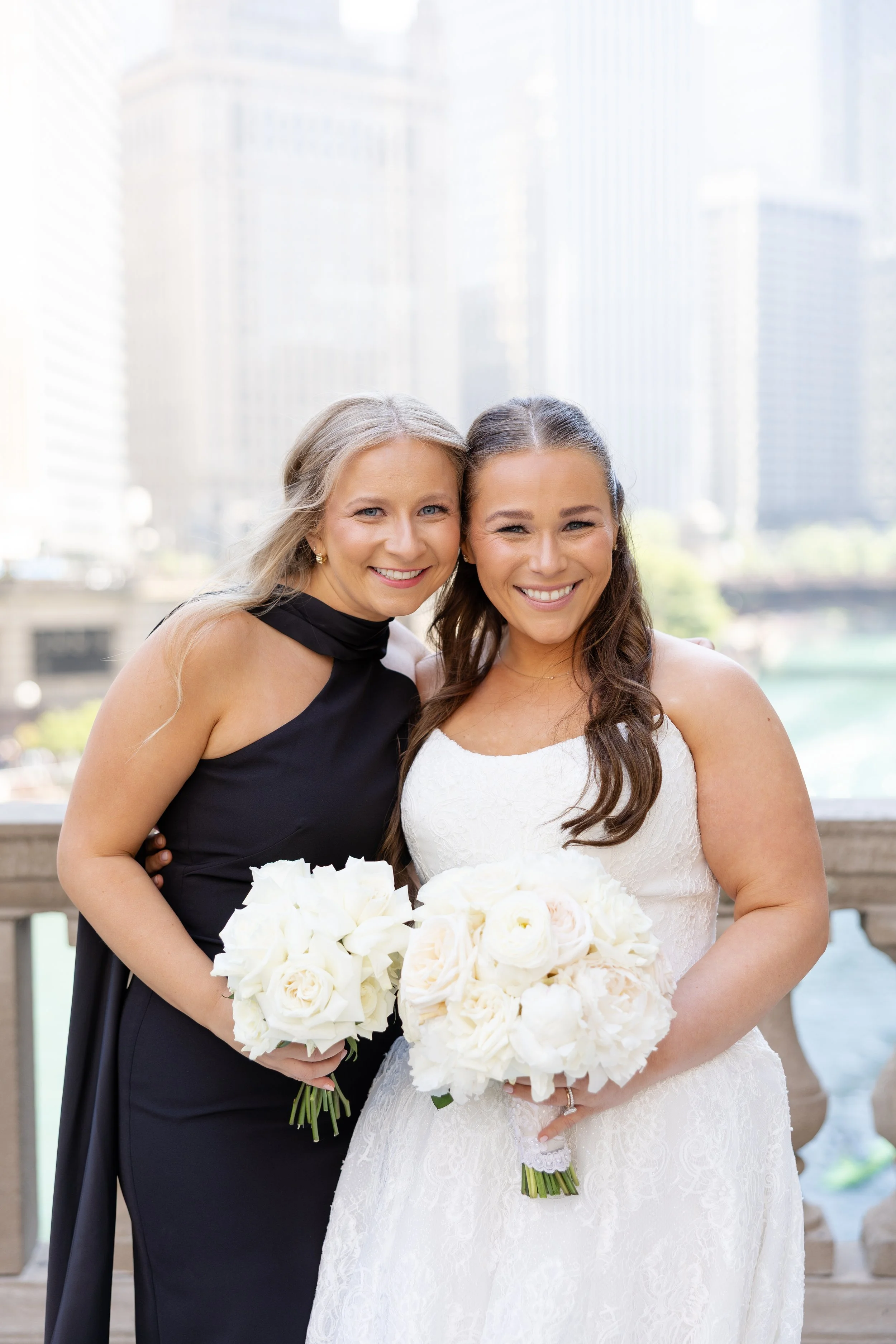 Two women, one in a black dress and the other in a white wedding gown, smiling and holding white bouquets in front of a city skyline with tall glass buildings.