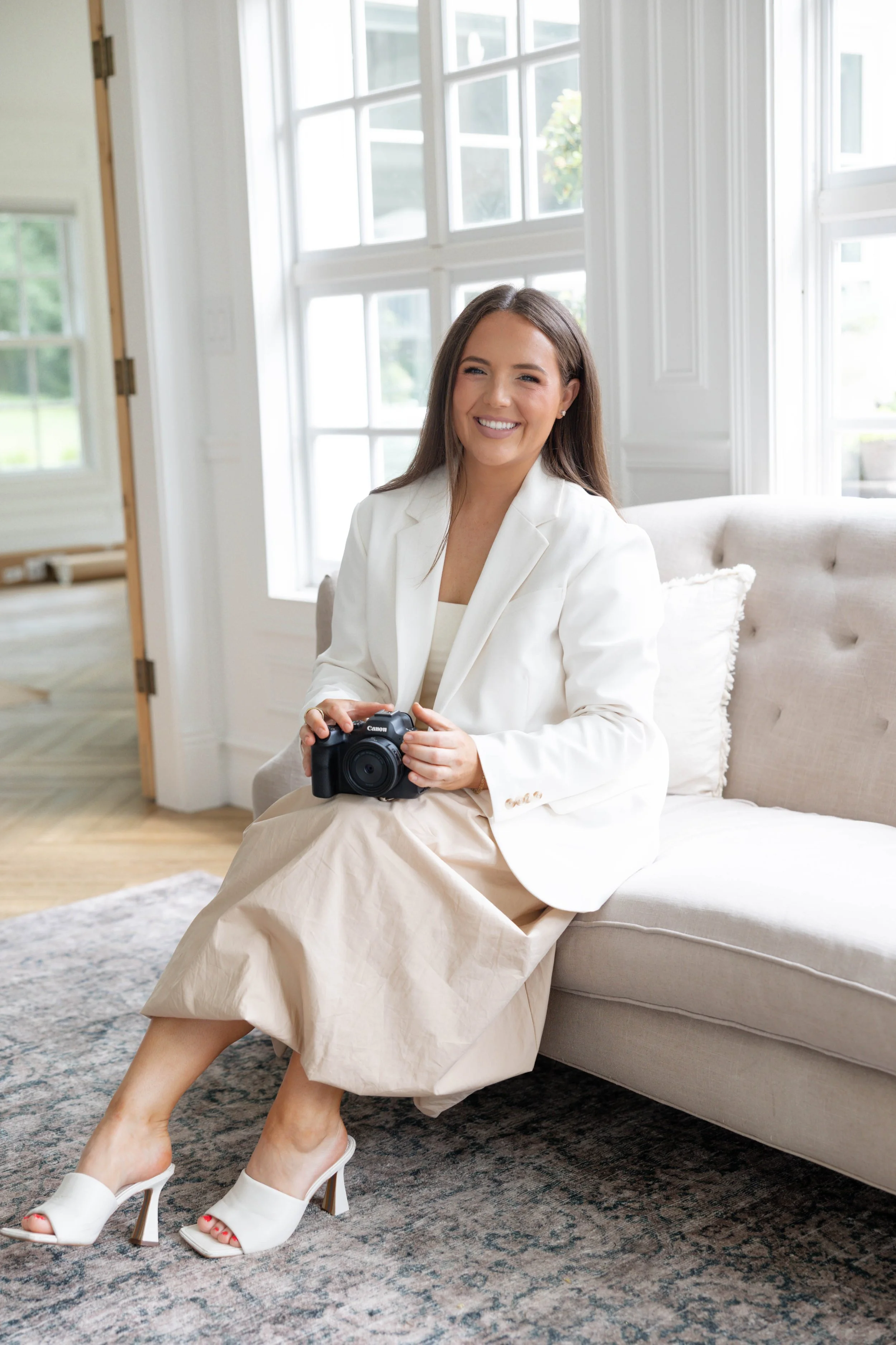 A woman sitting on a beige sofa in a bright room, holding a camera and smiling. She is dressed in a white blazer and a beige skirt, wearing white high heels. The room has large windows, white walls, and a gray patterned rug.