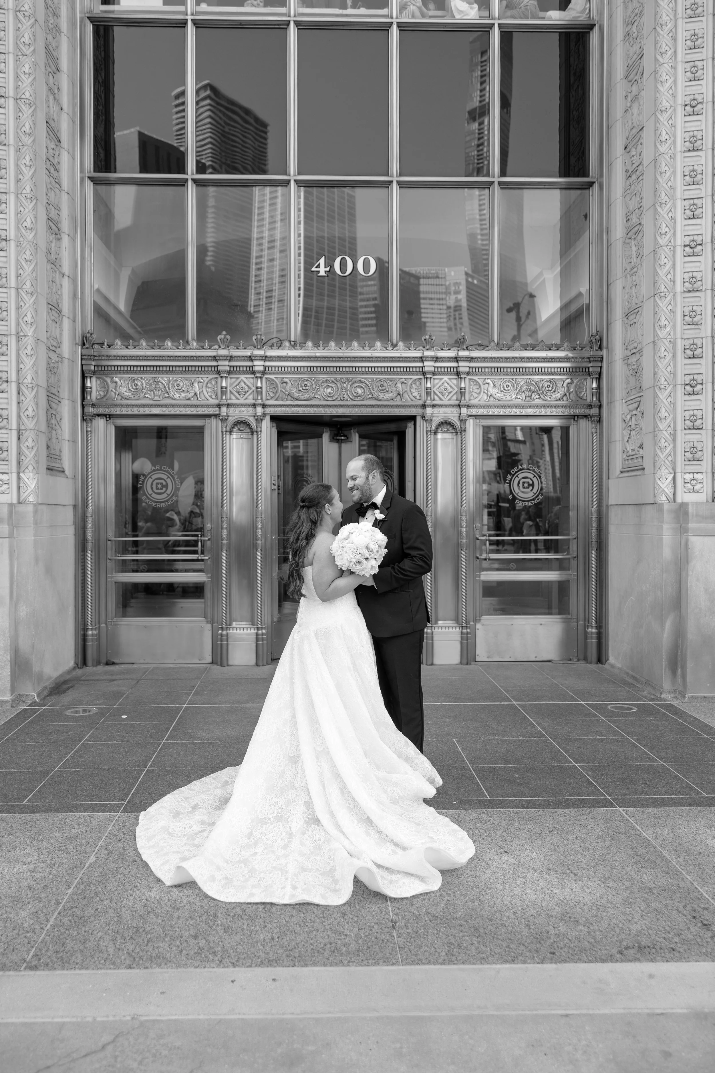 A bride and groom dressed in wedding attire stand close together in front of a large building entrance with the number 400 on the glass above. The bride wears a long, flowing lace wedding gown and holds a bouquet of flowers, while the groom is in a black tuxedo with a bow tie. They are smiling and appear to be in a joyful moment.