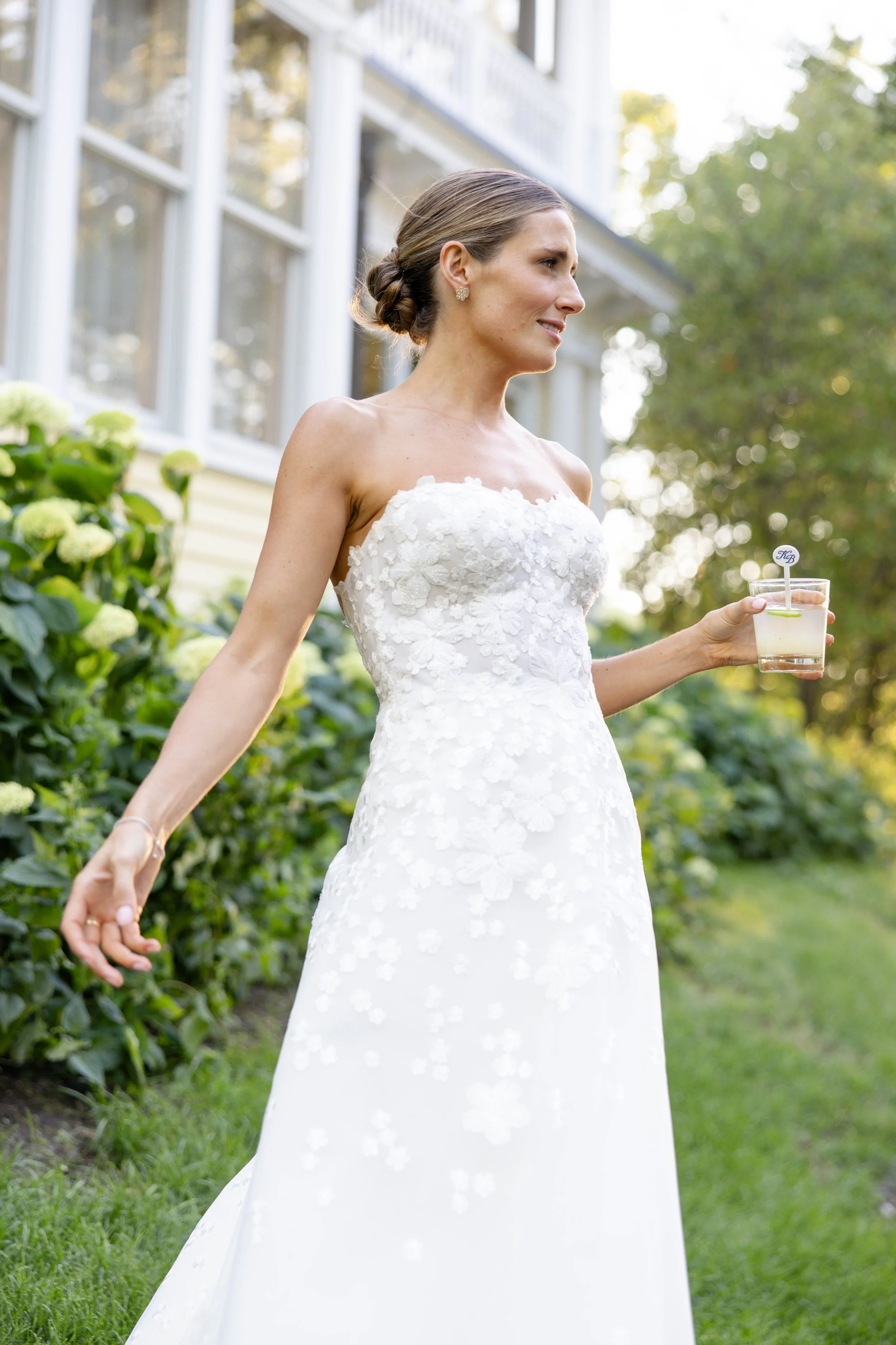 A woman in a white wedding dress standing outdoors, holding a drink with a lime wedge, with greenery and a house in the background.