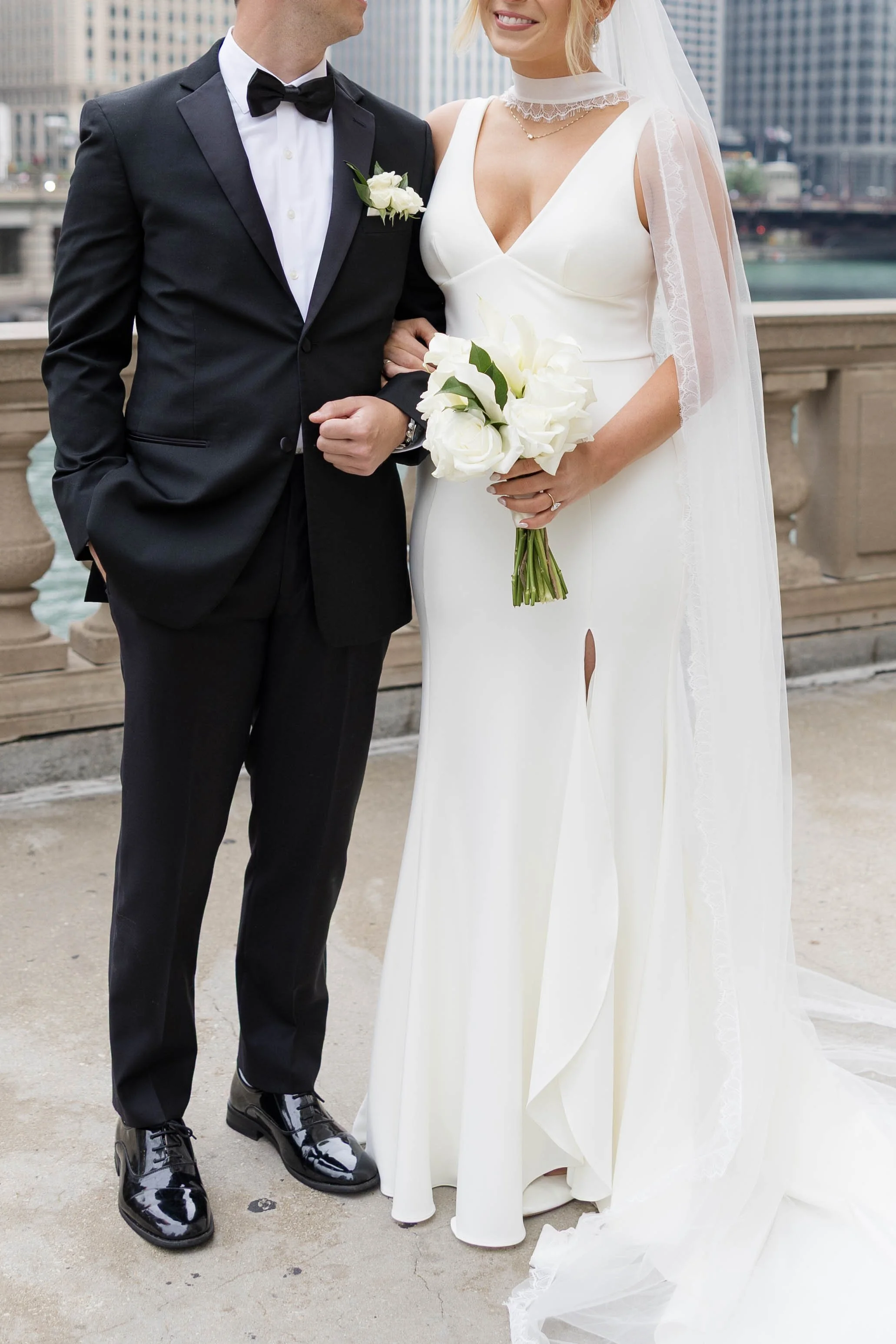 A bride and groom standing on a city rooftop. The groom wears a black tuxedo with a bow tie, and the bride wears a white wedding dress with lace details and a veil. The bride holds a bouquet of white flowers, and they are smiling at each other.