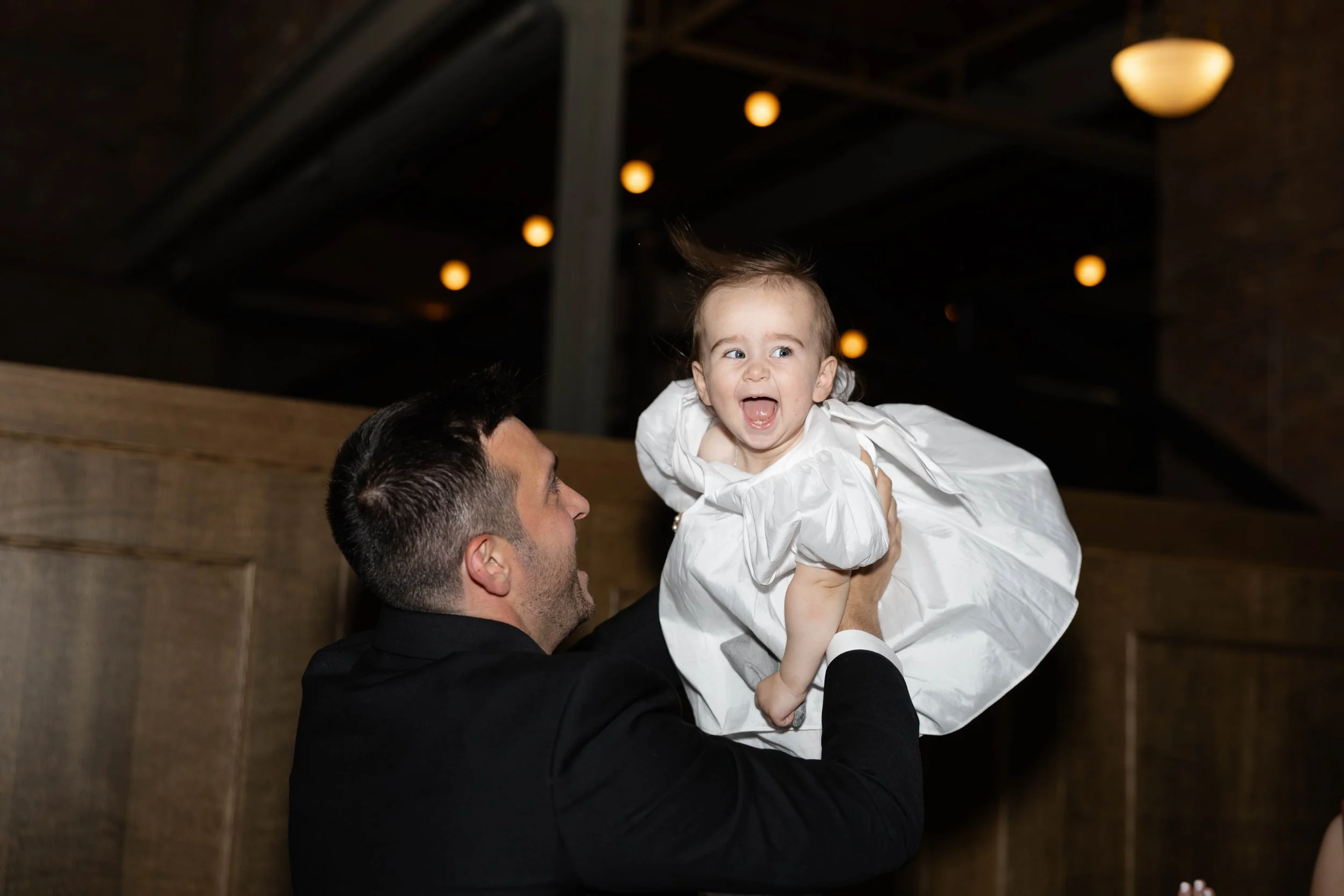 A man lifting a young girl in the air during a celebration, both smiling and looking happy. The girl is wearing a white dress, and the setting has warm lighting with a wooden backdrop.