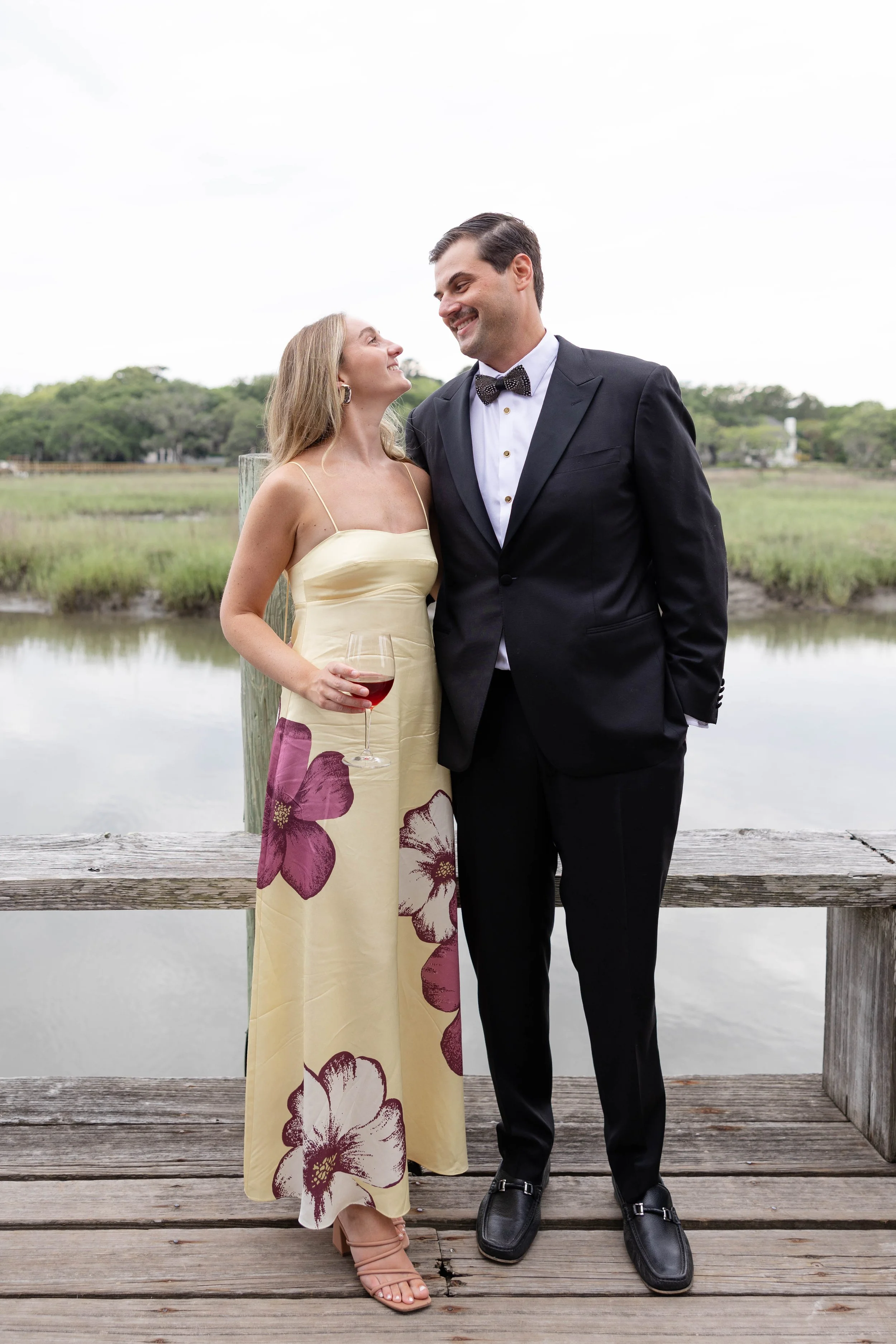 A couple dressed formally, embracing and smiling at each other on a wooden dock near a body of water with green vegetation in the background.