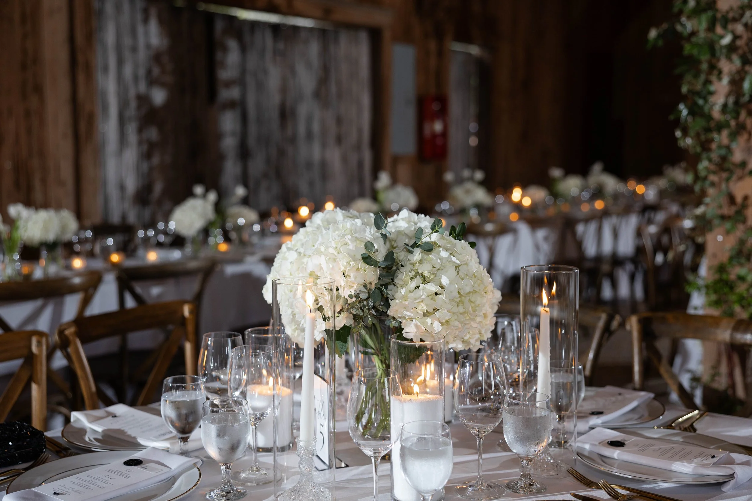 Elegant dining table setup with a large white floral centerpiece, surrounded by wine glasses, candles, and white napkins, in a rustic wooden venue with decor and chairs.