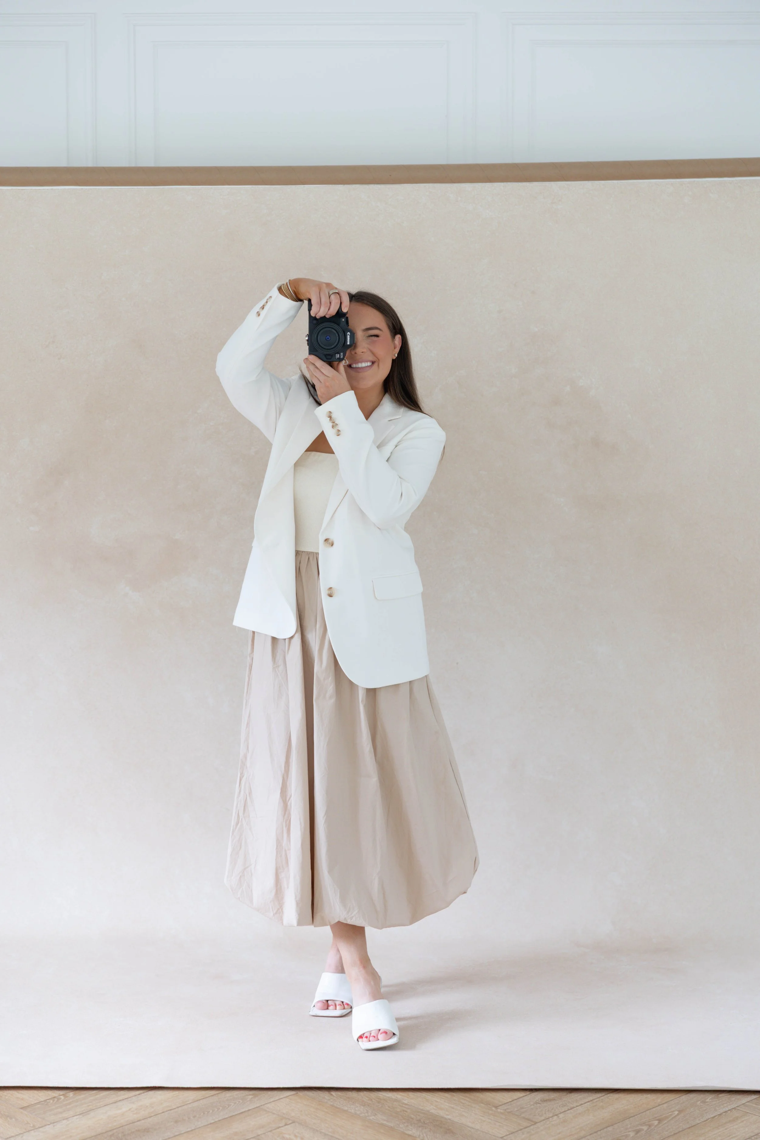 A woman taking a selfie with a camera, wearing a white blazer, beige wide pants, and white high-heeled shoes, standing against a beige textured wall.