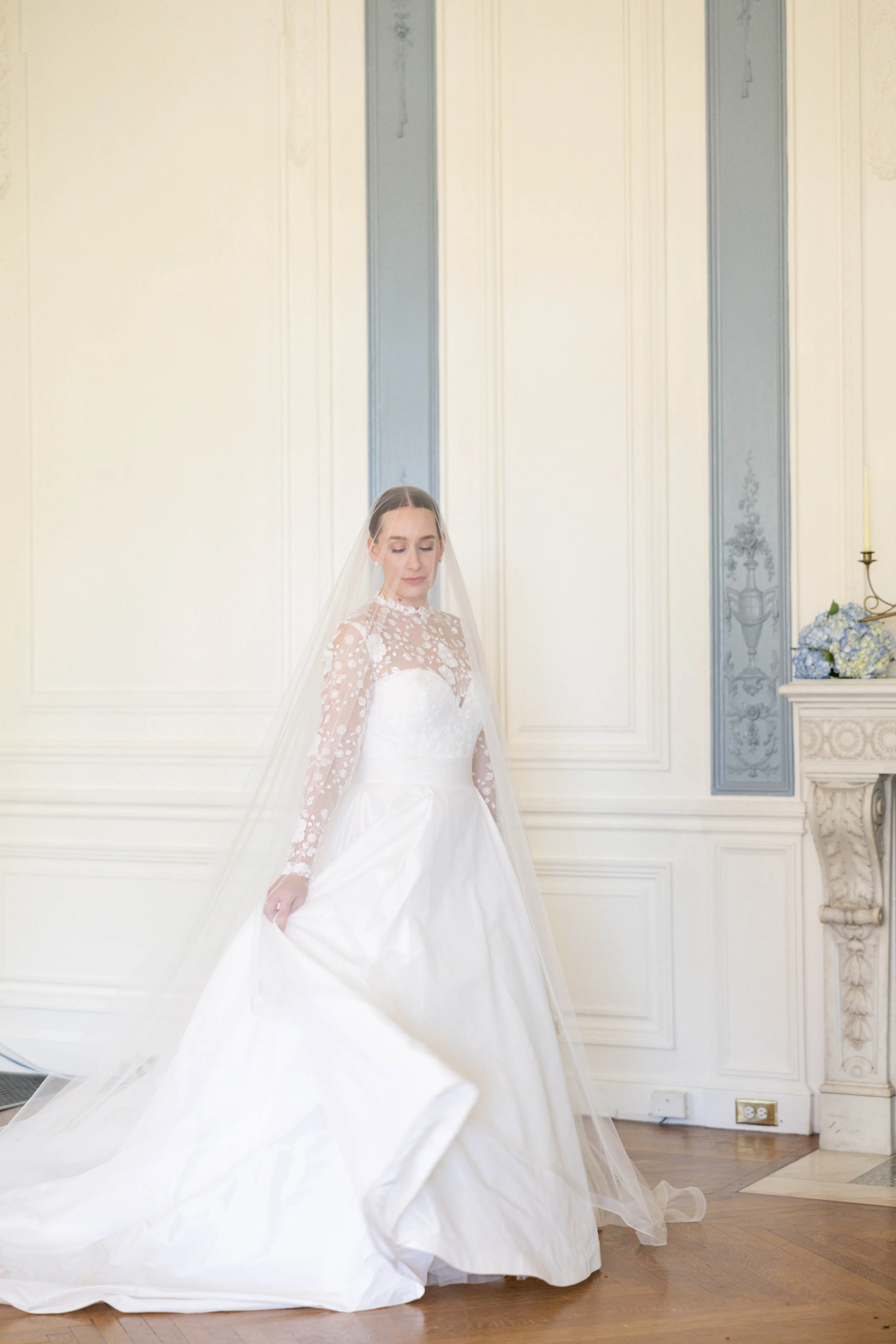 A bride in a white wedding dress with lace long sleeves, standing in a room with ornate wall paneling, holding up the hem of her dress with one hand and looking down, with a veil over her face.
