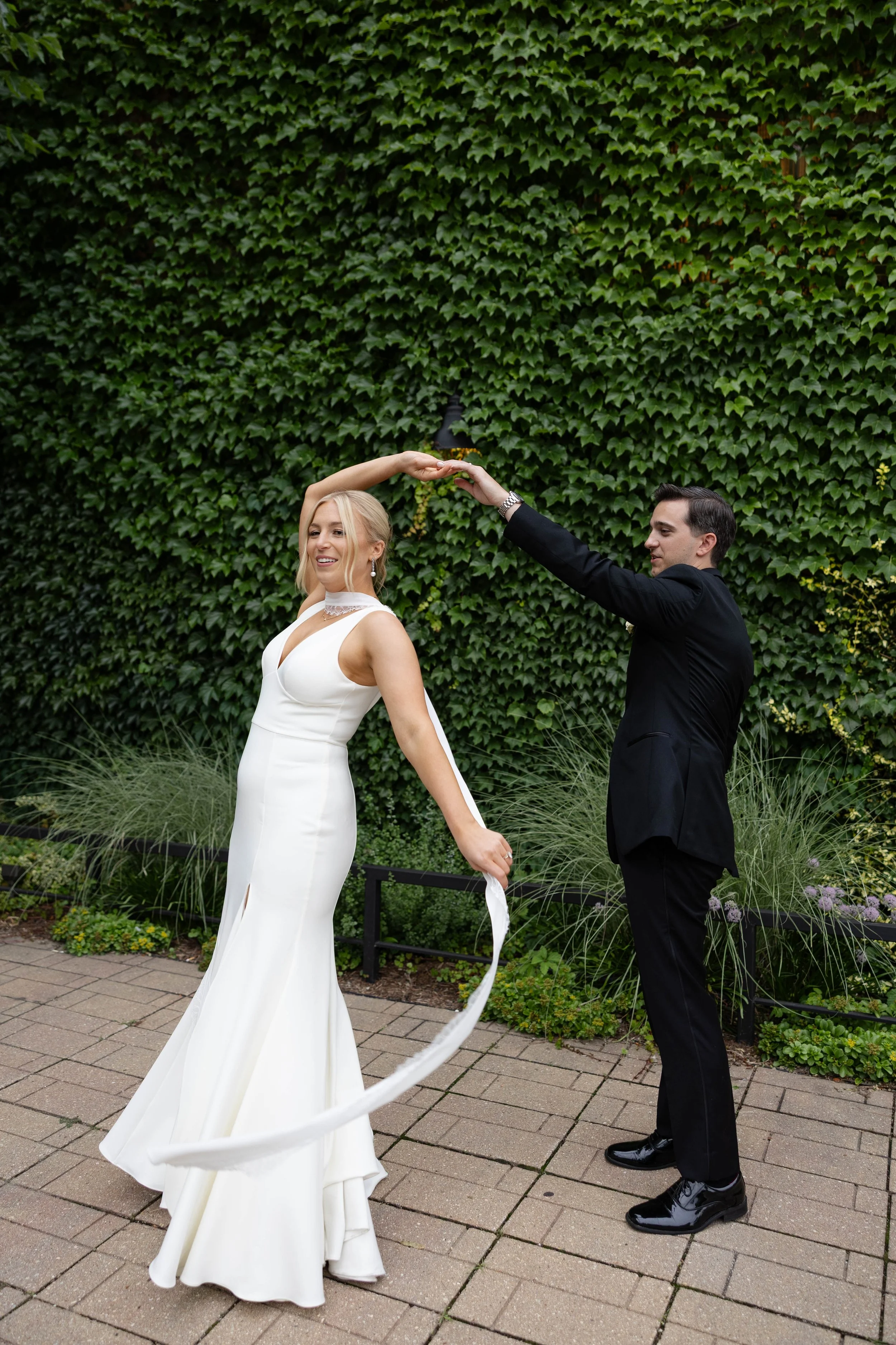 A woman in a white wedding dress and a man in a black suit dancing outdoors with a green ivy-covered wall in the background.