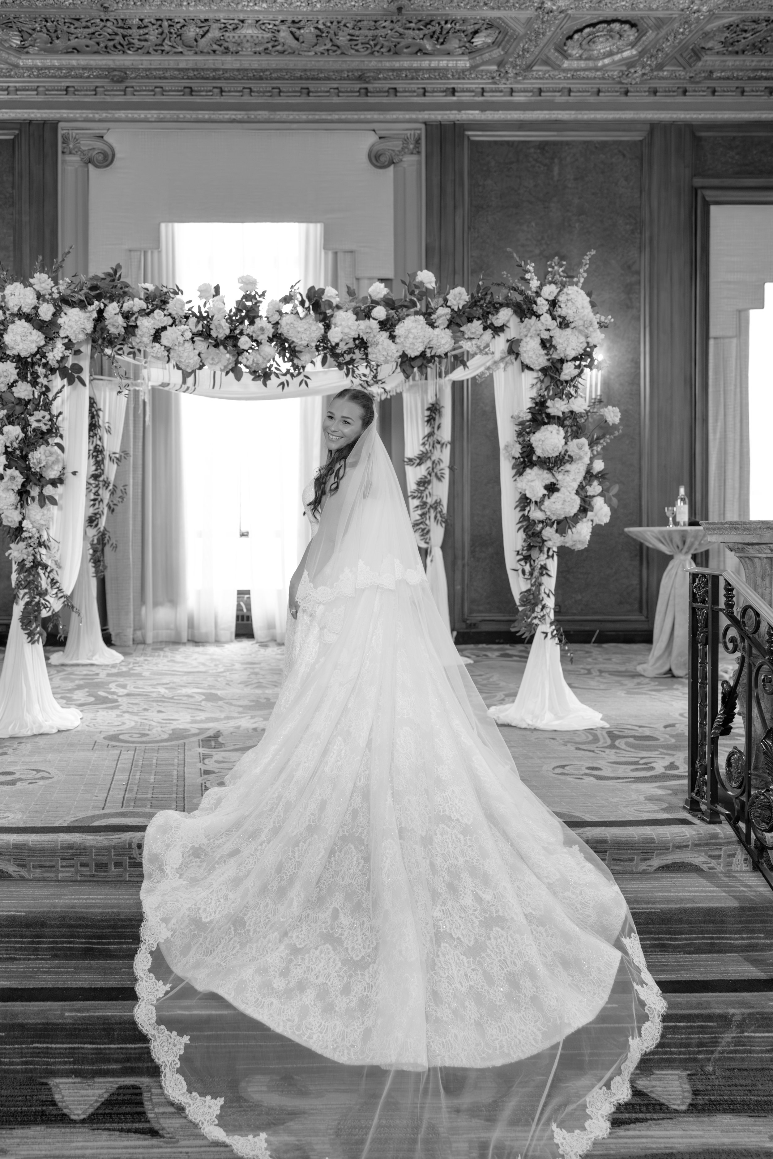 A bride in a wedding dress standing in a decorated indoor venue with floral arrangements and drapery, smiling toward the camera.