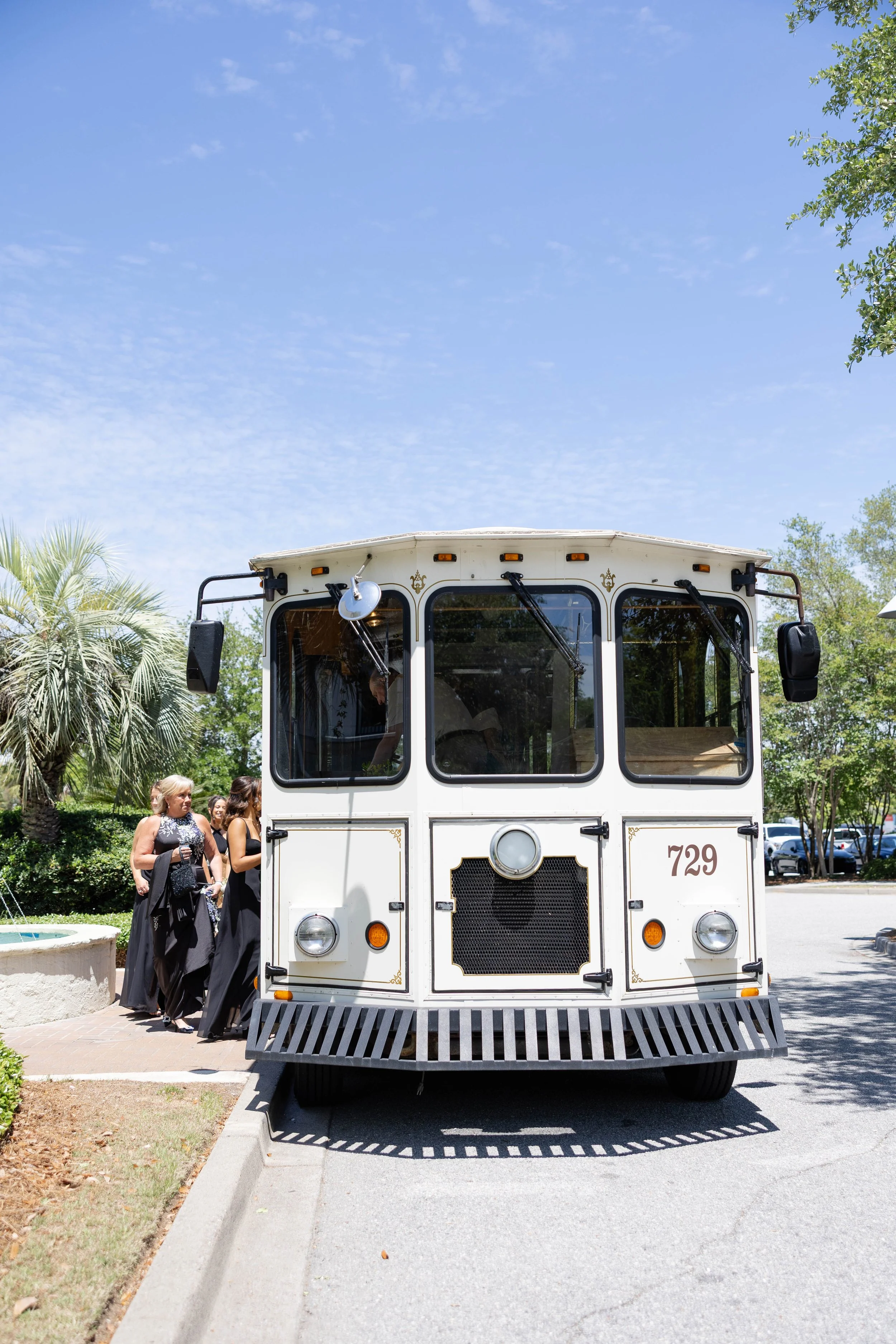 A white trolley with gold accents and the number 729 on the front, parked on a street with a group of women dressed in black walking nearby. Trees are visible in the background and a clear blue sky overhead.