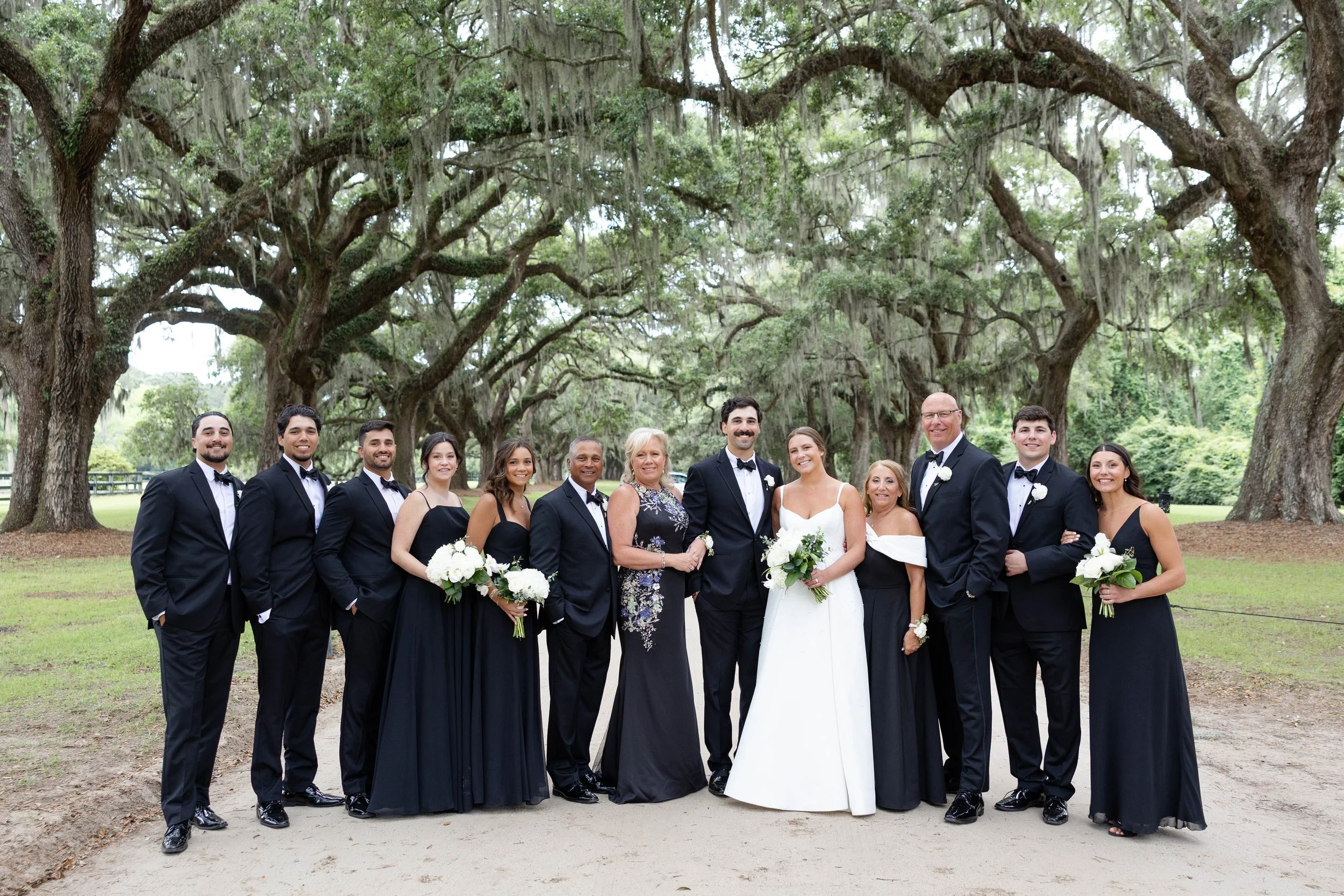 A group of people, including brides and grooms, dressed in wedding attire, standing outdoors under large oak trees with Spanish moss, celebrating a wedding.