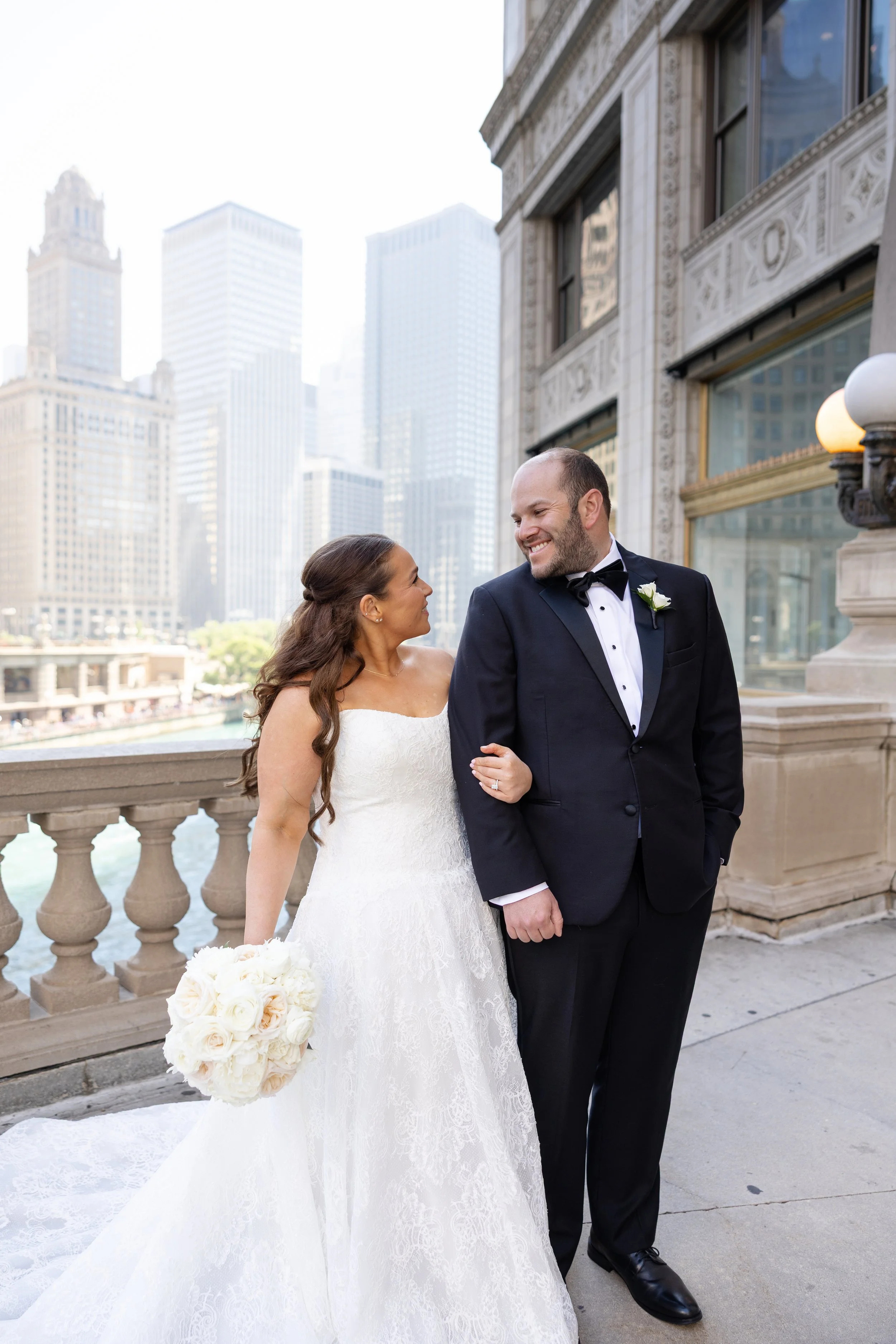 A bride and groom smiling at each other on a city bridge, with tall buildings in the background. The bride is holding a bouquet of white flowers, and the groom is wearing a black tuxedo with a bow tie and a white boutonniere.